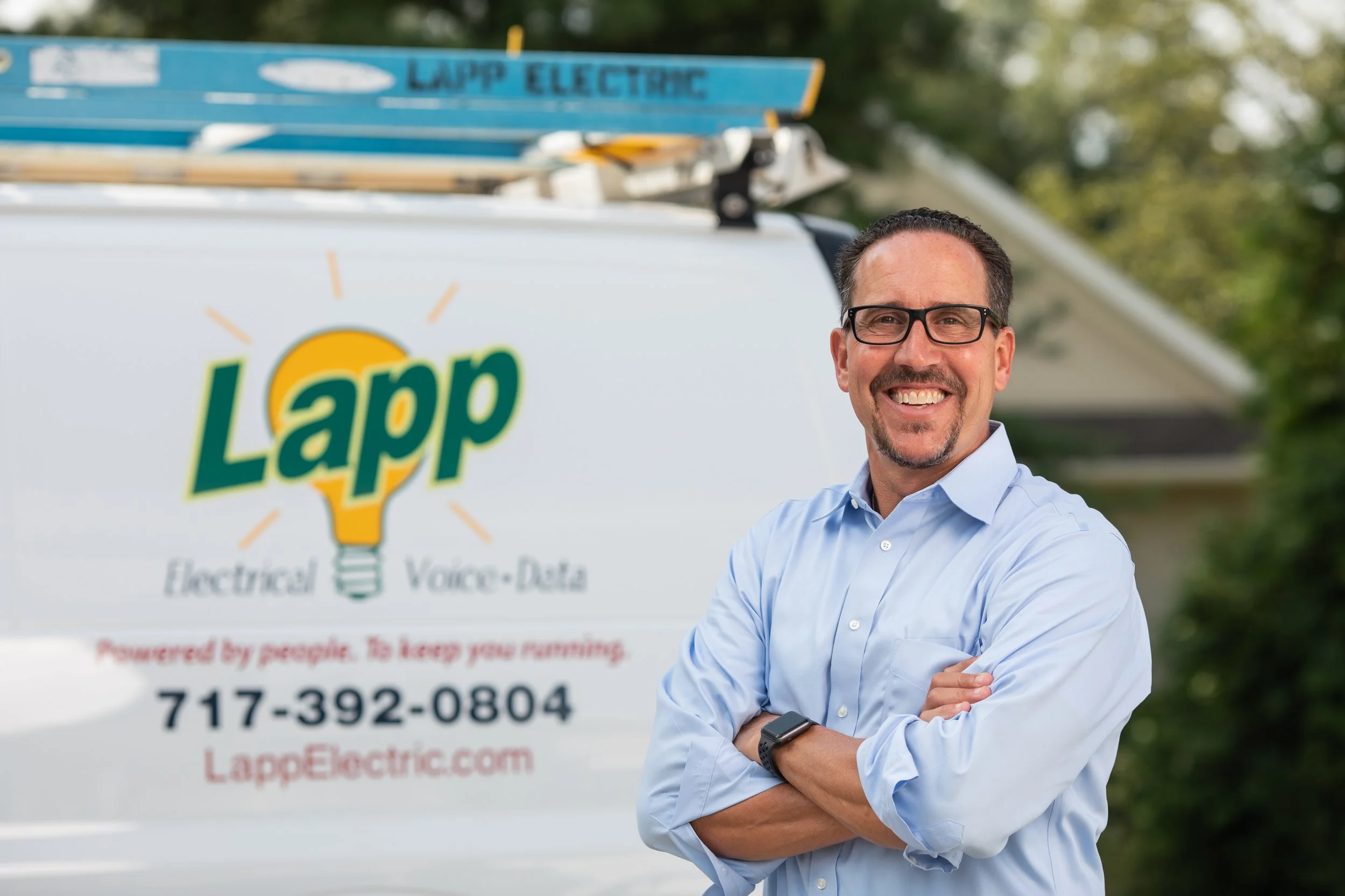 A man with glasses and a beard smiling with arms crossed, standing outdoors in front of a Lapp Electric service vehicle, which has the company's logo and contact information.