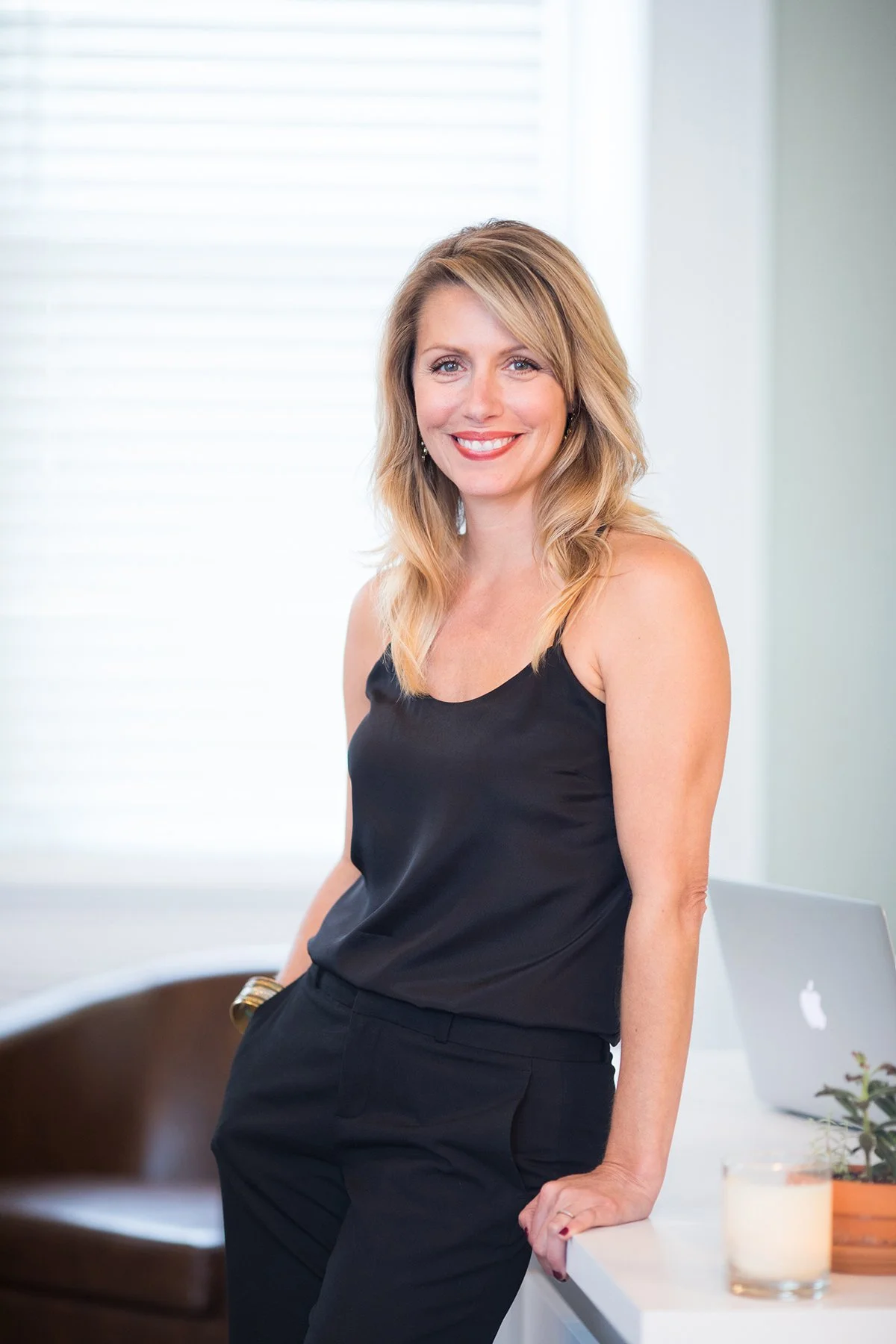 A woman with blonde hair in a black sleeveless top and black pants smiling while standing in a bright room near a white table with a laptop, candle, and plant.