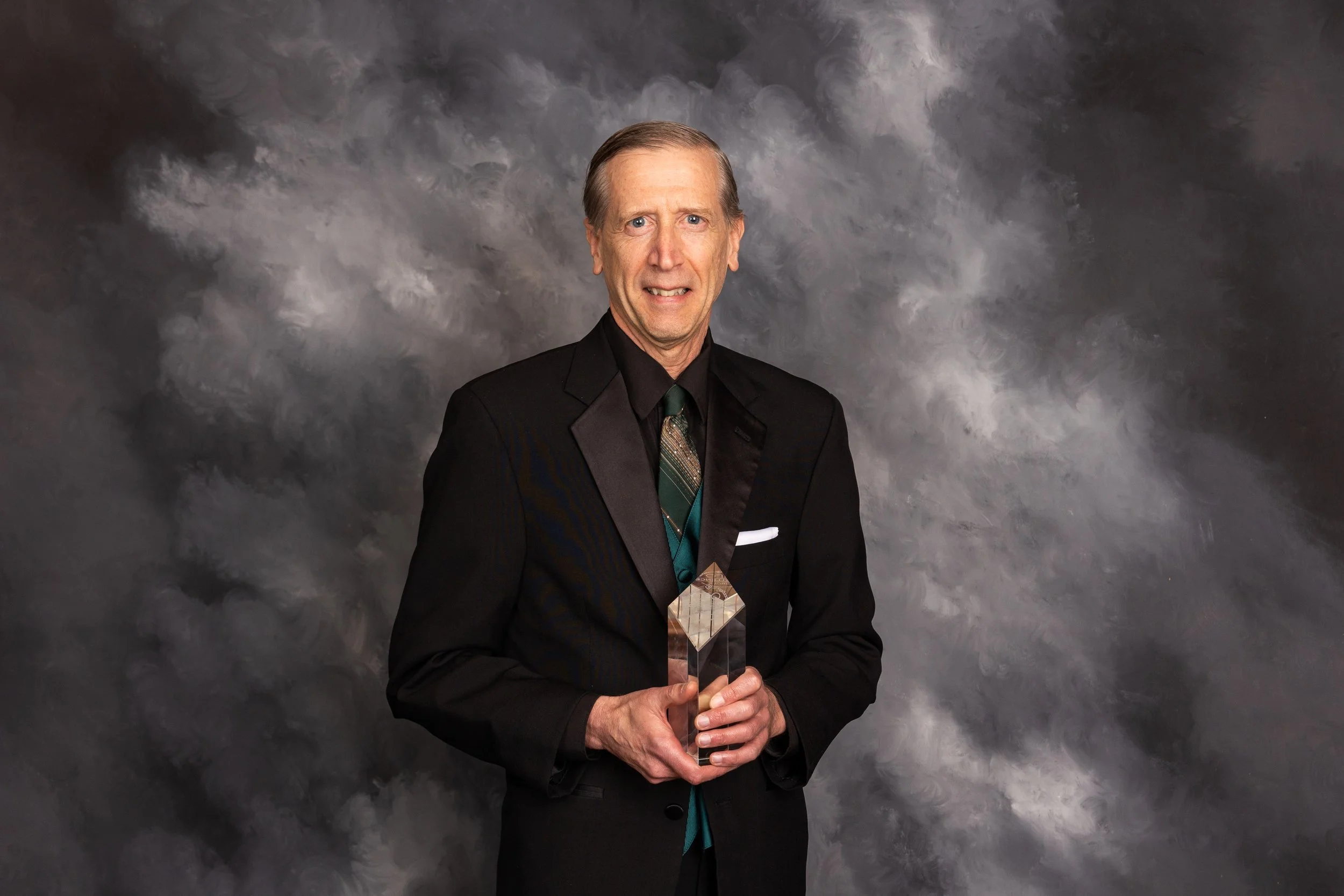 A middle-aged man in a black suit with a patterned tie, holding a glass award, standing against a gray cloudy background, with a confused or distressed expression on his face.