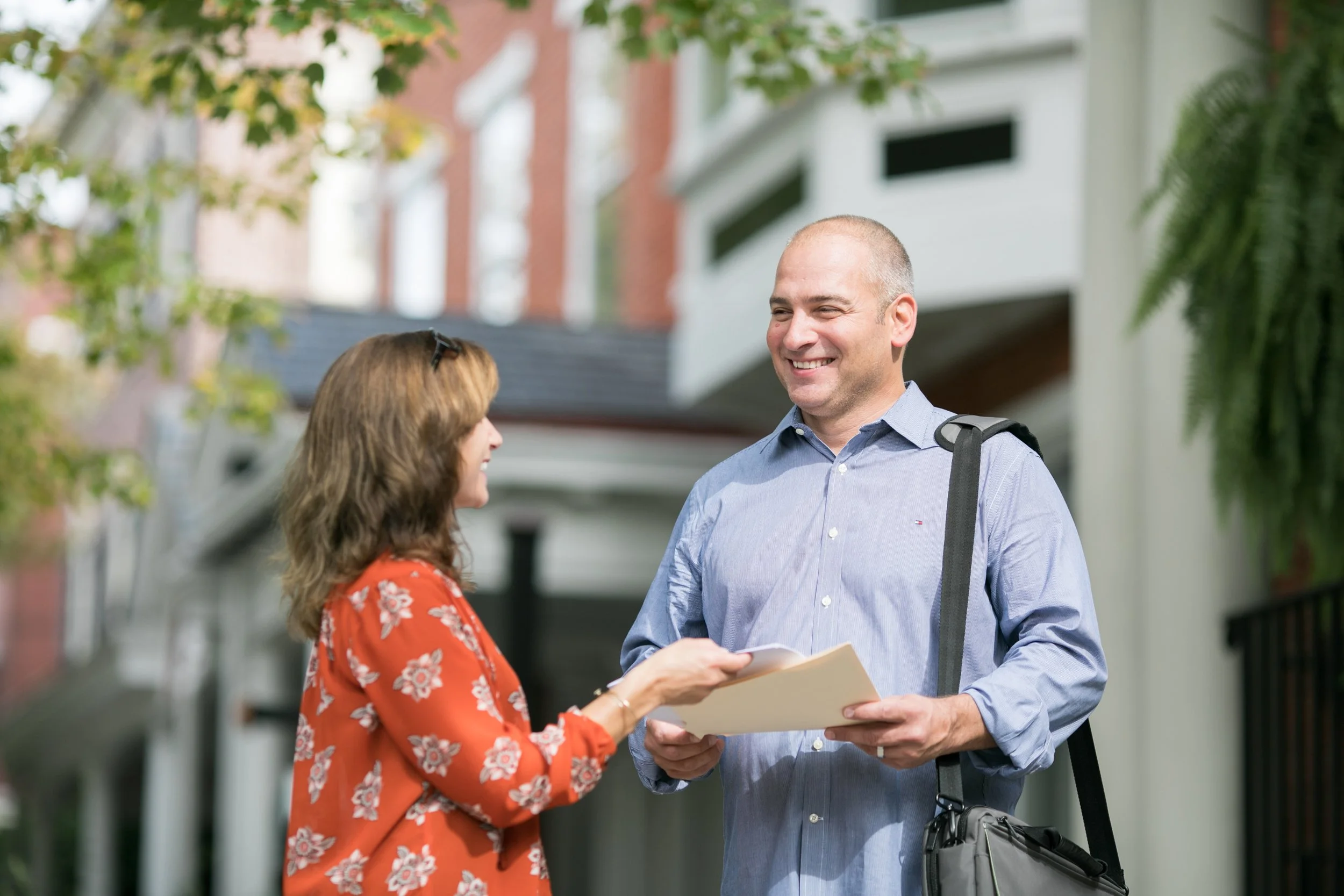 A man and woman are talking outside in front of houses on a sunny day. The woman is holding papers, and the man is smiling and carrying a bag over his shoulder.