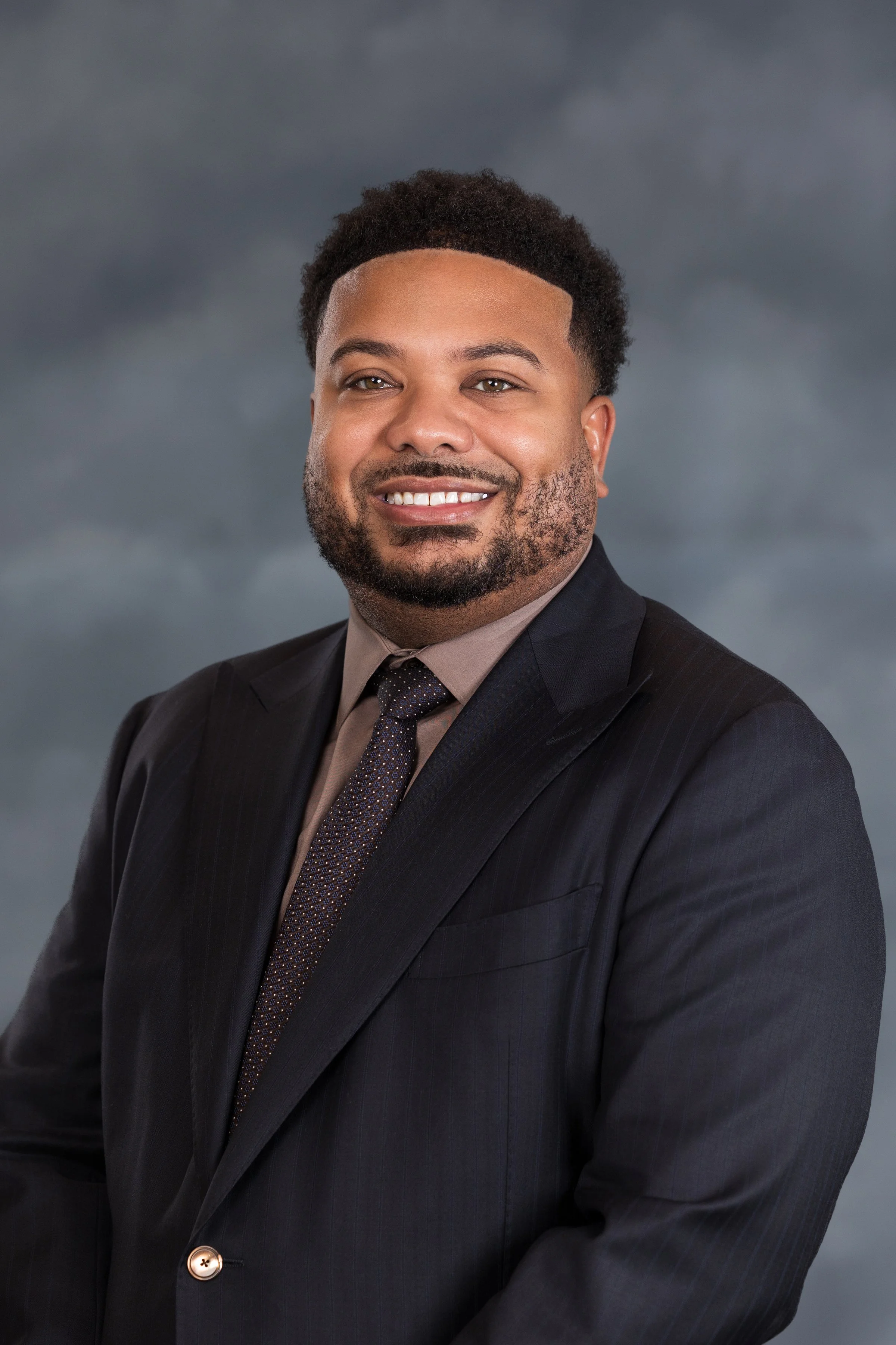 Professional portrait of a smiling man wearing a dark suit, light shirt, and patterned tie, standing against a cloudy sky background.