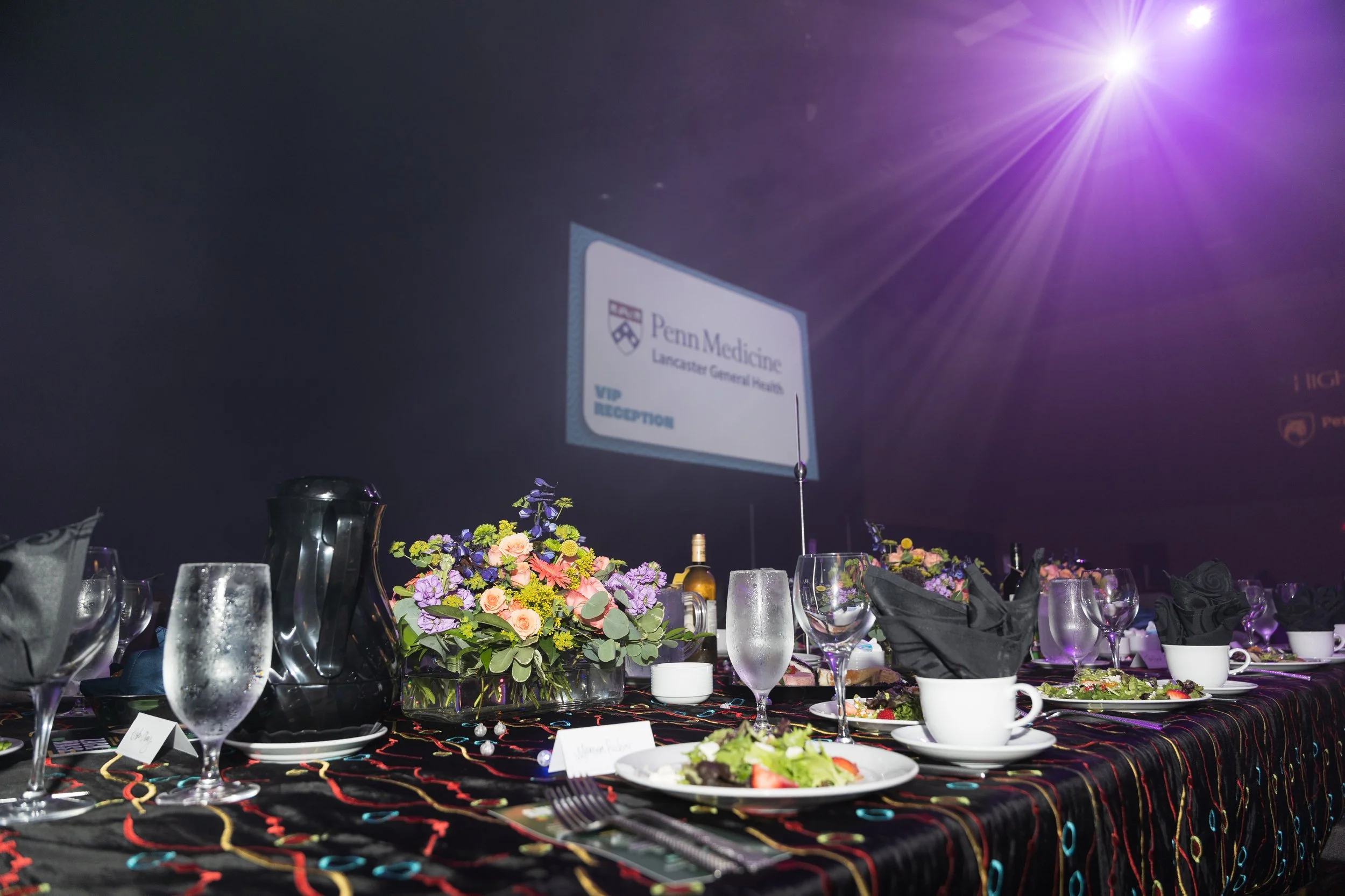 A decorated banquet table with floral centerpieces, glassware, plates, and black napkins, set up for an event at Penn Medicine Lancaster General Health, with a large screen displaying the Penn Medicine logo in the background and purple lighting overh