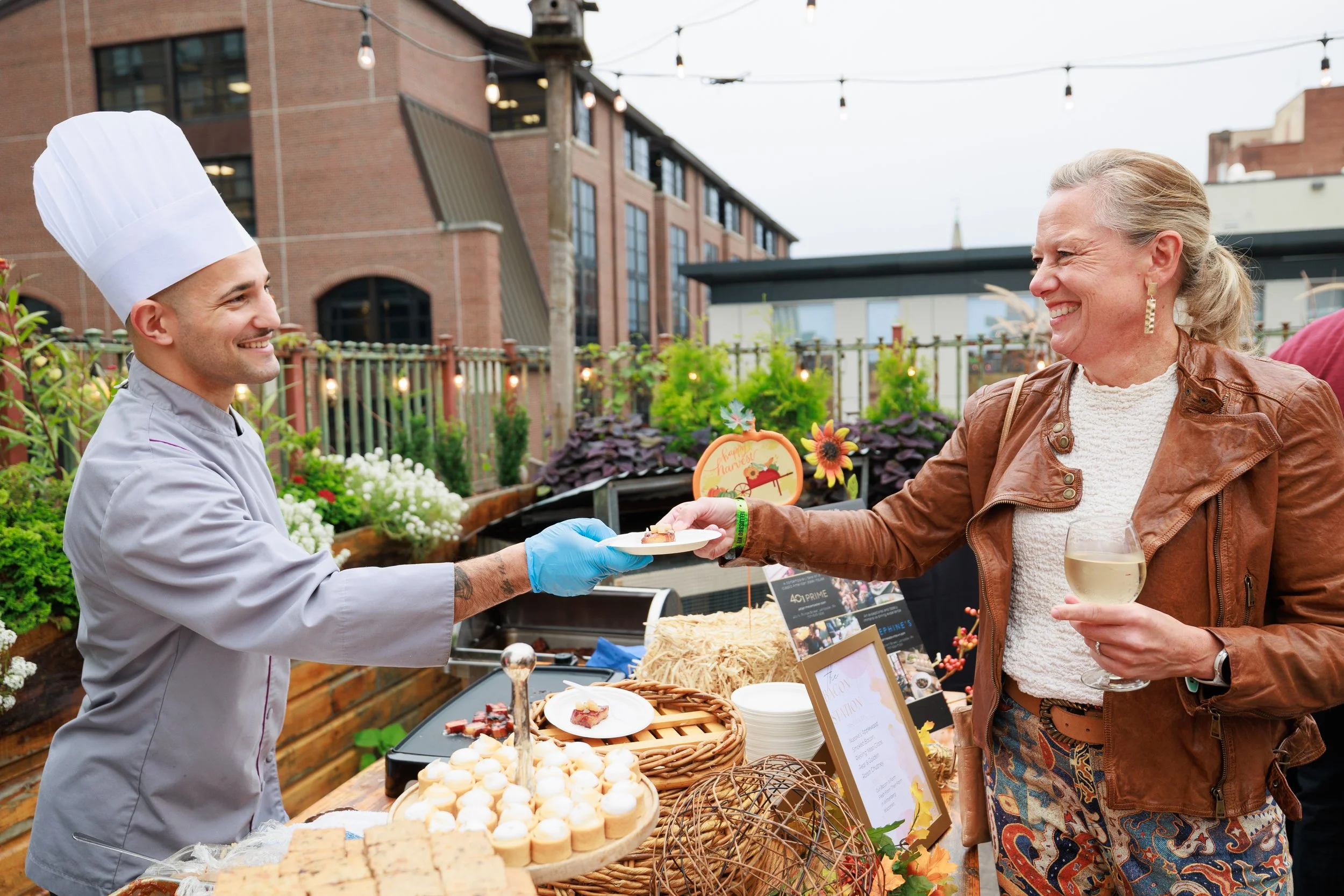 A smiling chef handing a small plate of food to a woman holding a glass of white wine at an outdoor event with decorated food table and string lights.
