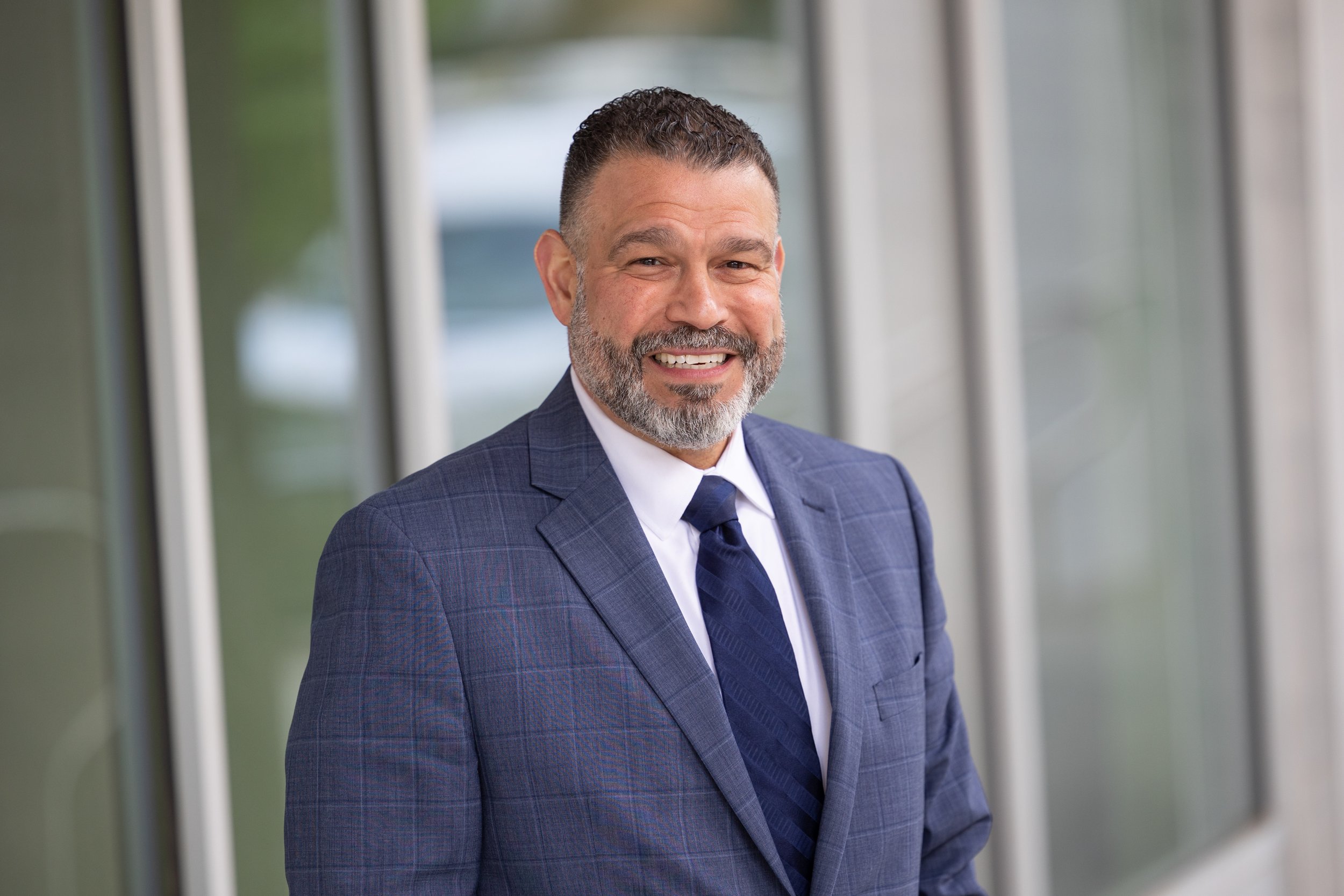 Smiling man in a blue plaid suit with a white shirt and dark blue tie standing in front of a window.