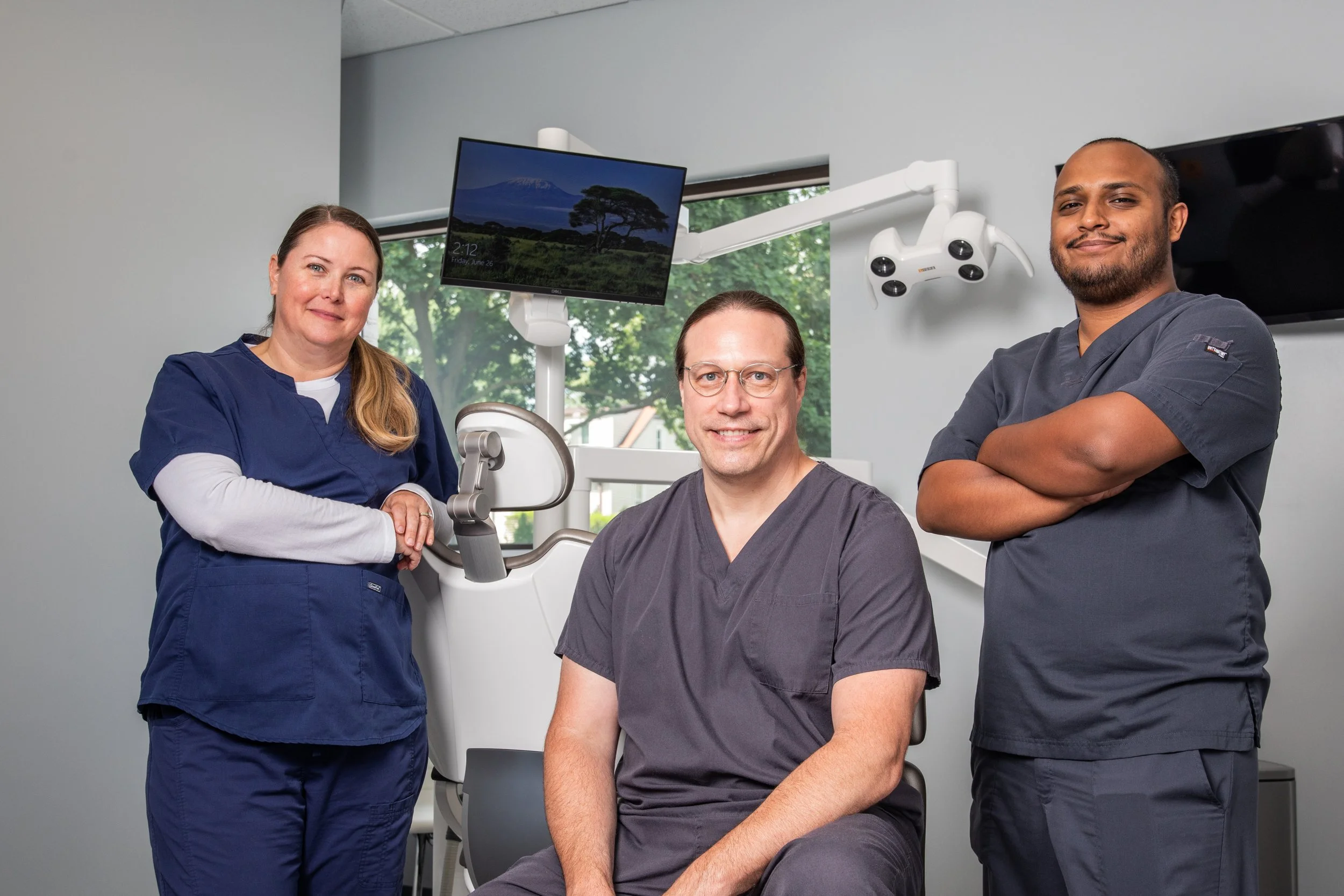 Three medical professionals in scrubs in a clinical setting with dental or surgical equipment and a monitor in the background.