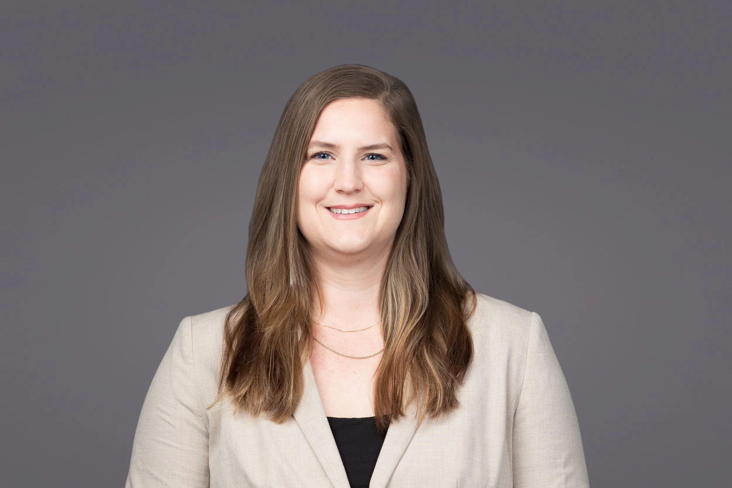 A portrait photograph of a woman with long, wavy brown hair, blue eyes, and a light beige blazer over a black top, smiling against a plain gray background.