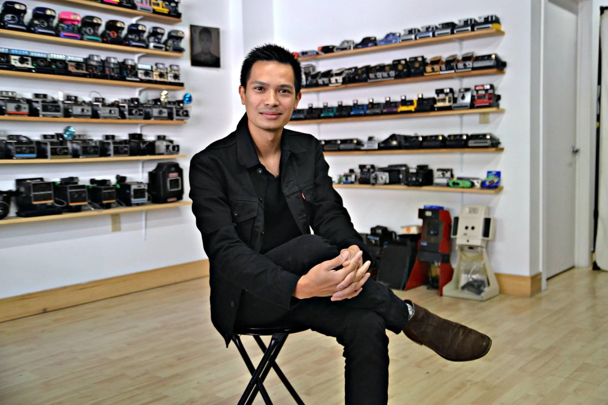 A man sitting on a black folding chair in a room with shelves filled with vintage cameras and cameras. He is wearing a black jacket and black pants, with one leg crossed over the other.
