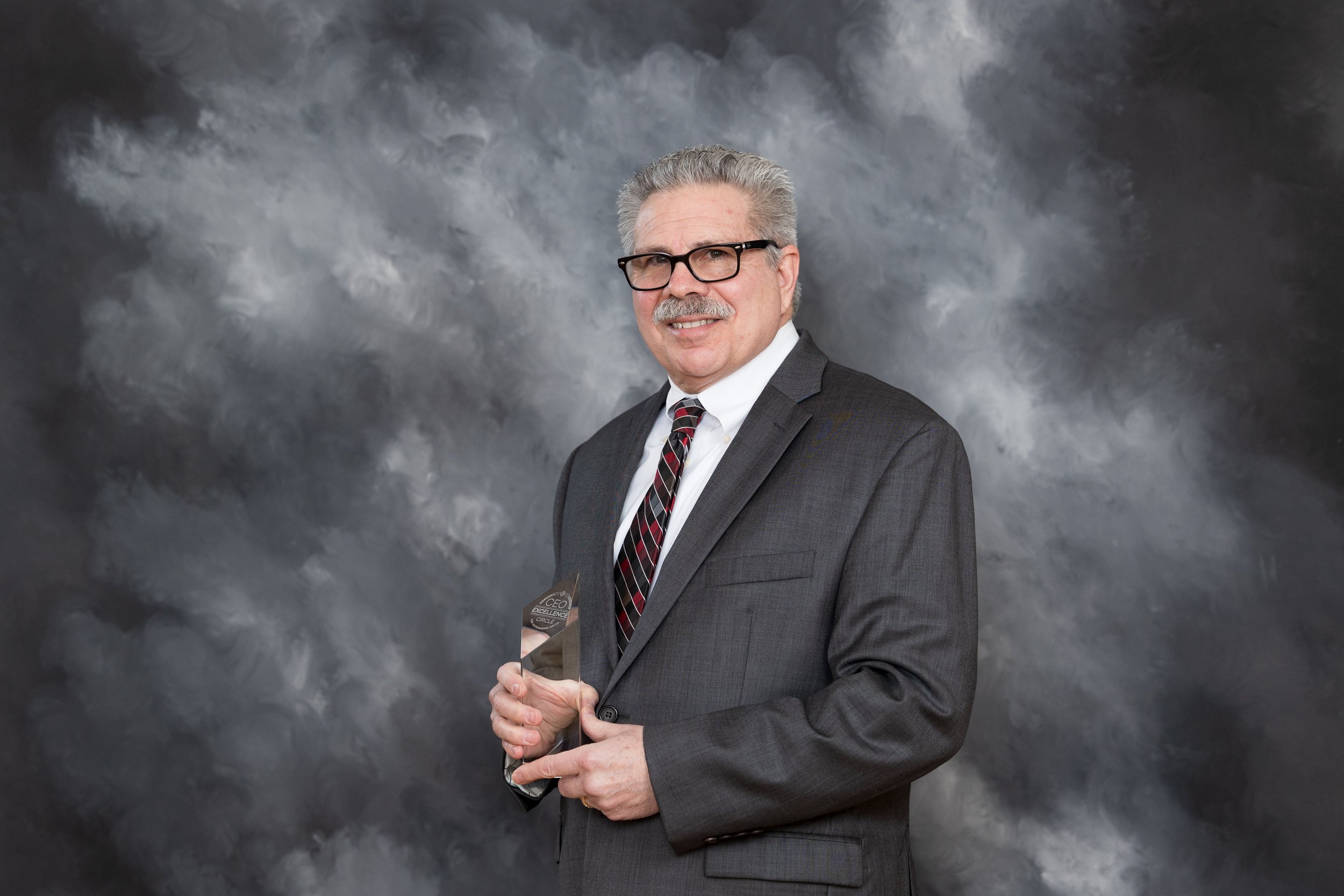 An older man with gray hair and glasses in a gray suit, white shirt, and striped tie, holding an award while smiling at the camera in front of a gray cloudy background.