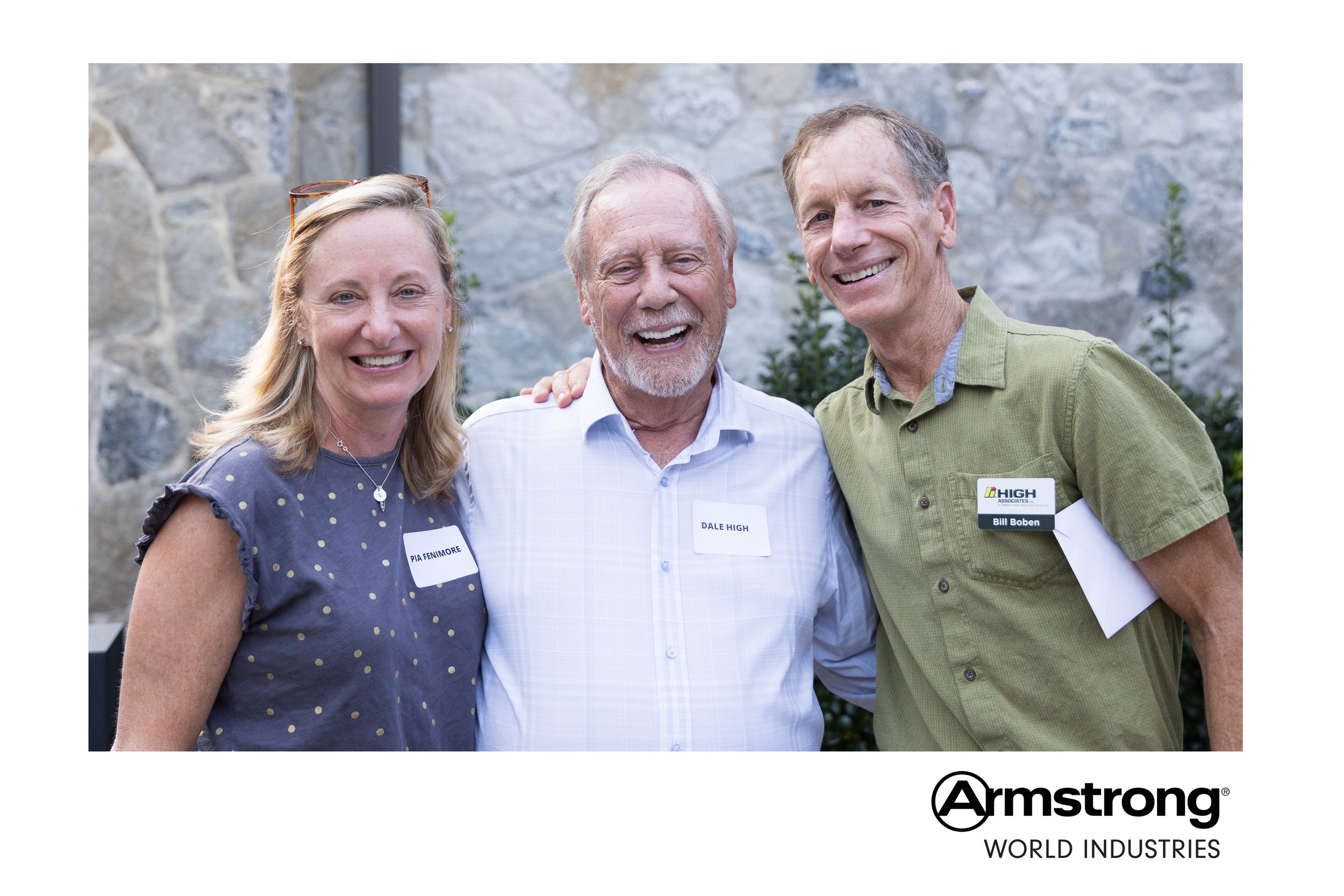 Three smiling people at an outdoor event, wearing name tags, with a stone wall and greenery in the background.
