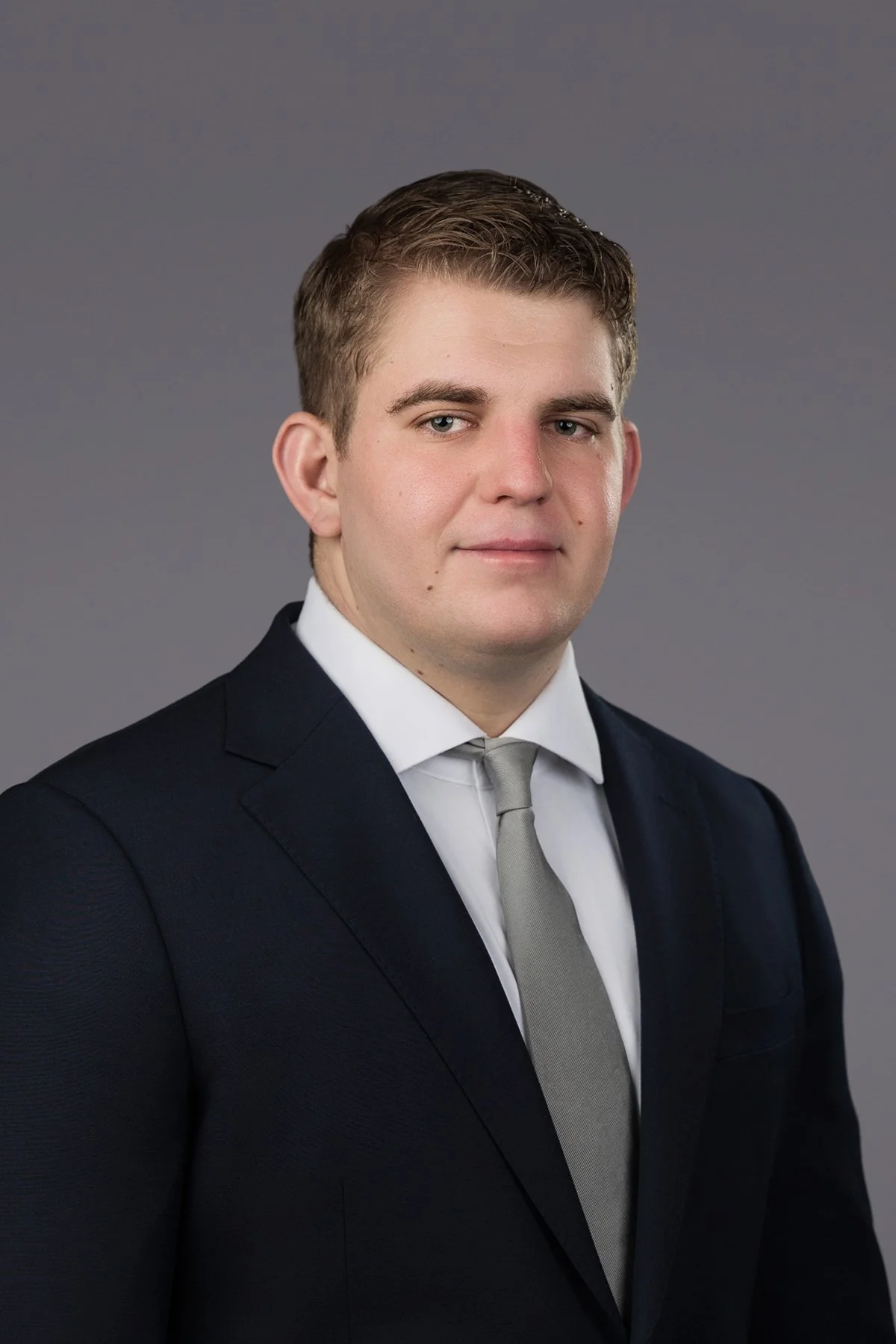 A young man dressed in a business suit, white shirt, and gray tie, posing against a plain gray background.