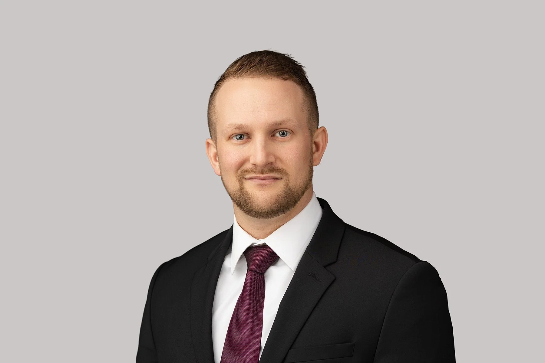 Professional portrait of a man in a black suit, white shirt, and maroon tie, against a plain light gray background.