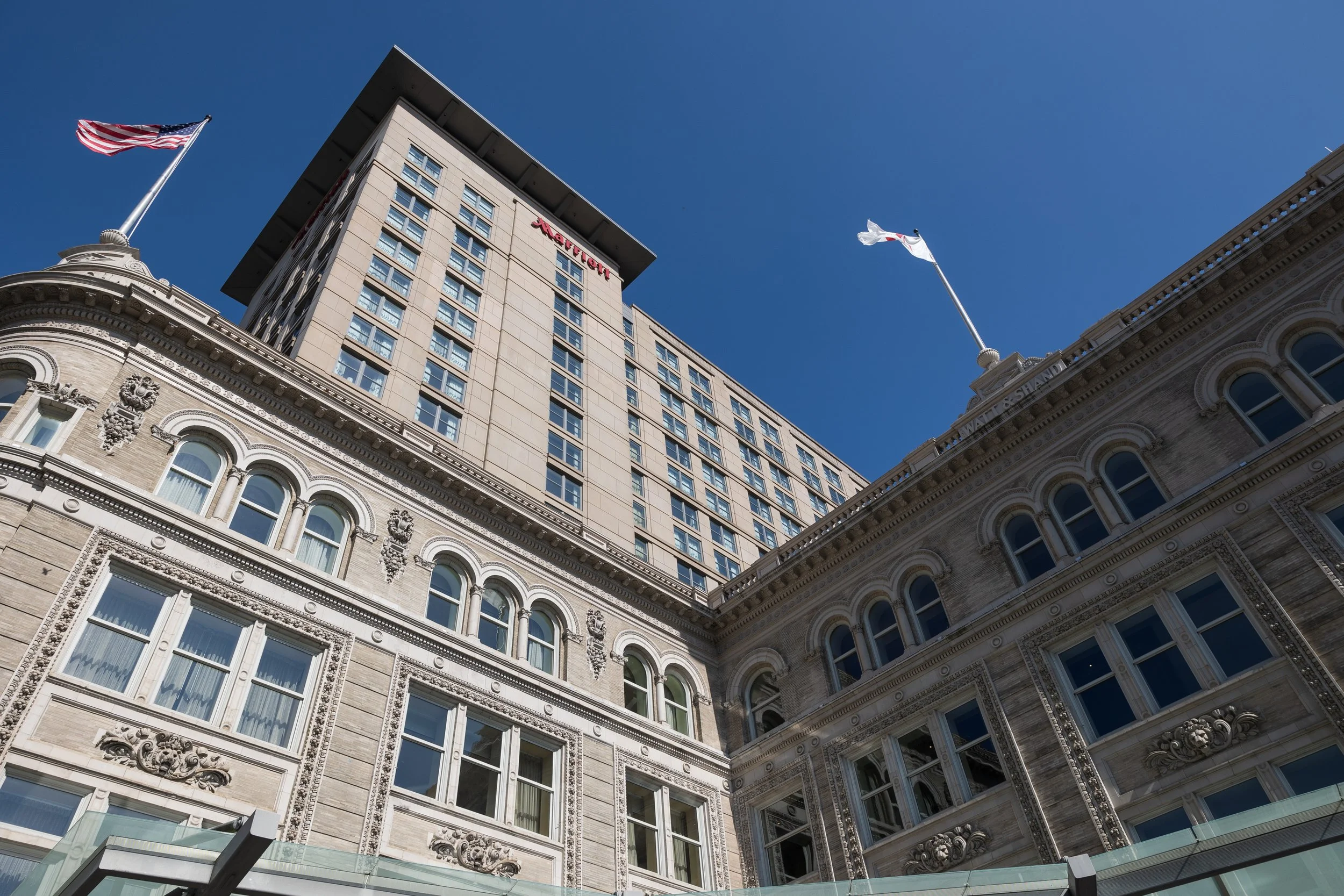 Low-angle view of historic and modern buildings under a clear blue sky, with American flags flying.