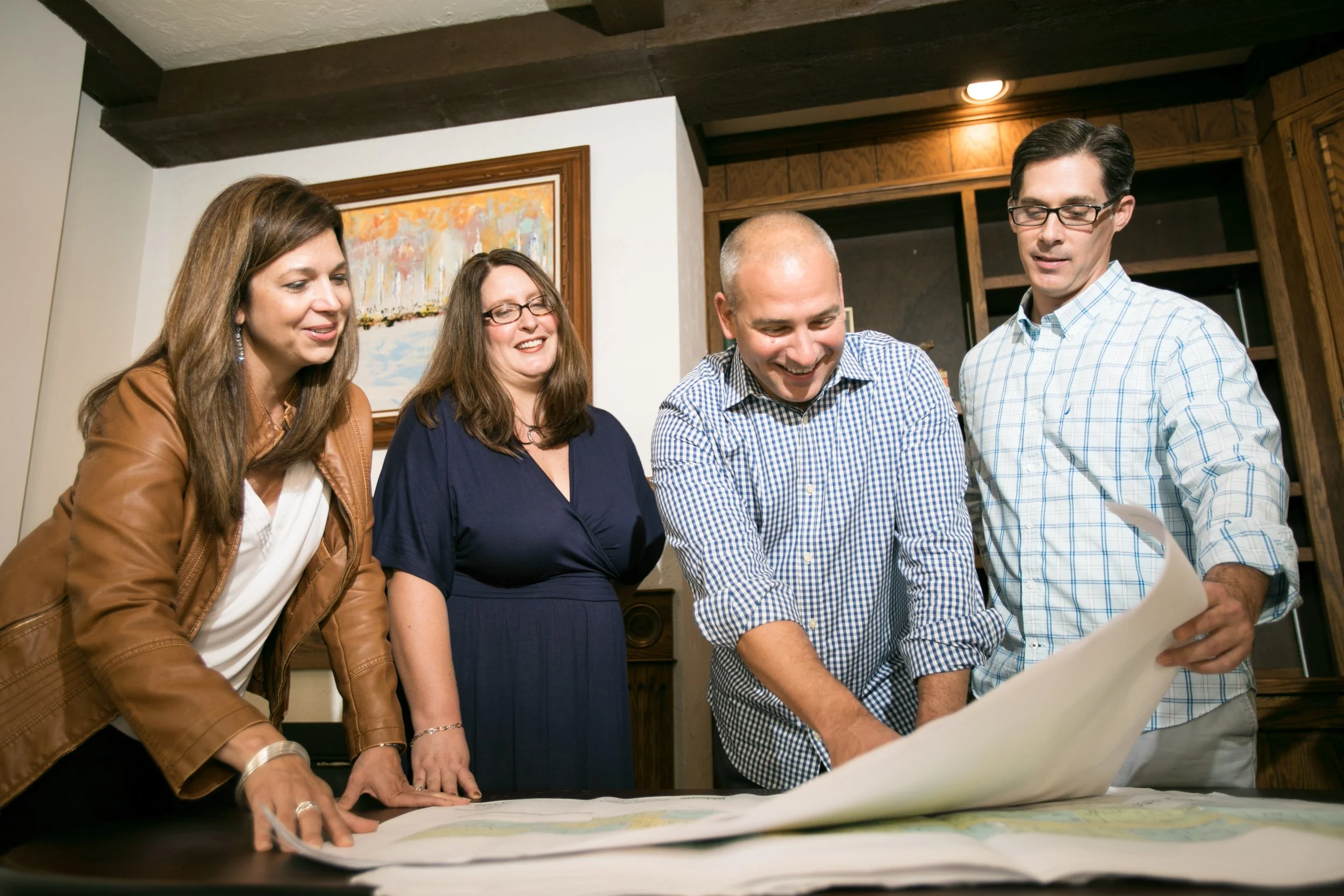 Four adults, two women and two men, gathered around a table looking at a large map, smiling and engaging in discussion.