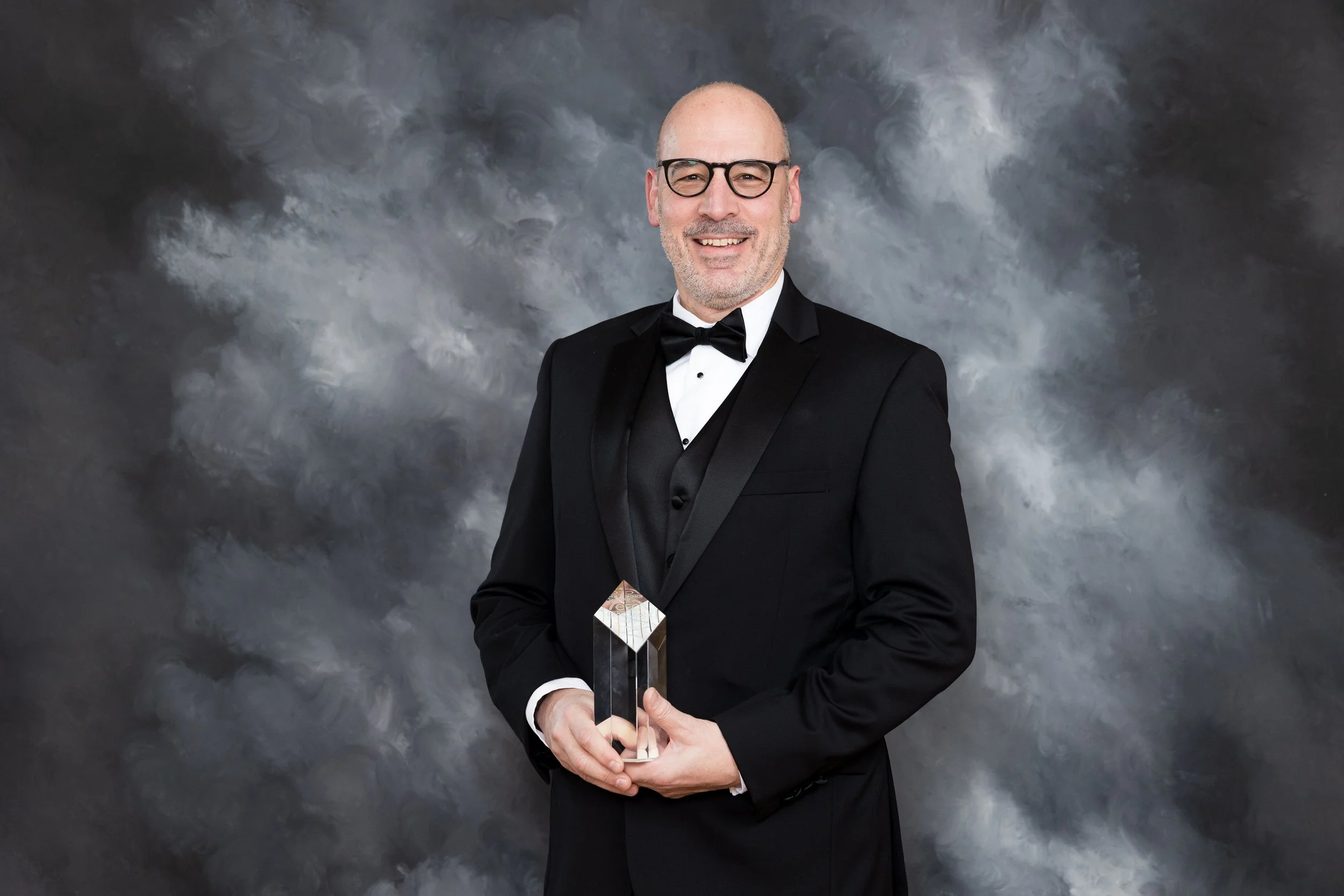 A man in a black tuxedo with a bow tie and glasses is smiling and holding a glass award, standing in front of a gray abstract backdrop.