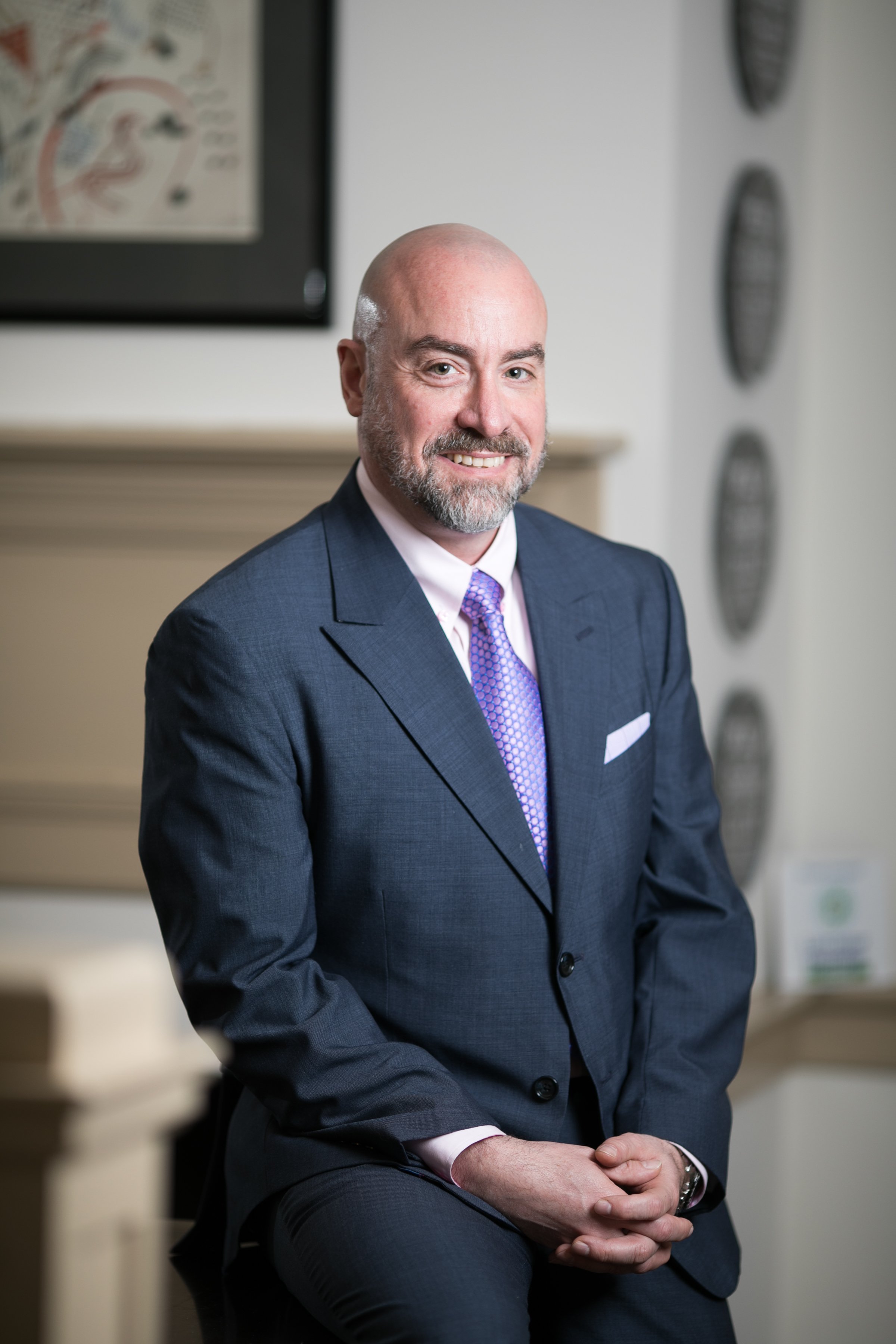 Professional man with a bald head and beard, wearing a dark suit, white shirt, and purple tie, sitting indoors with a friendly smile.