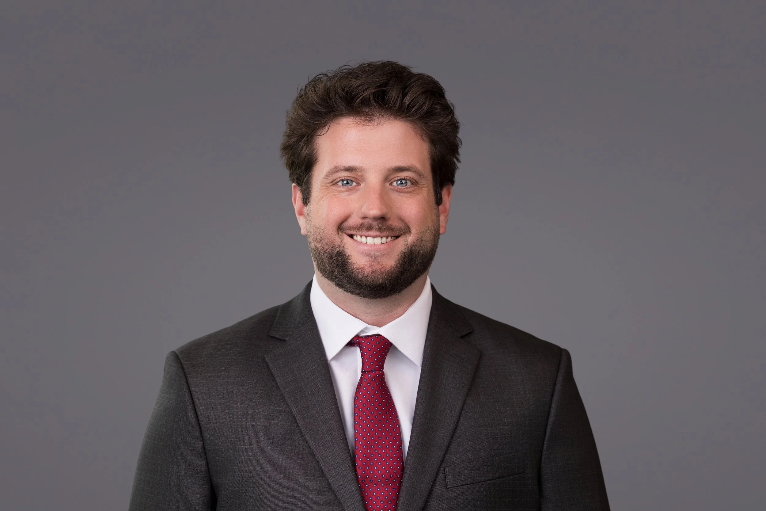 A man with brown hair and a beard, wearing a dark suit, white shirt, and red dotted tie, smiling against a plain gray background.
