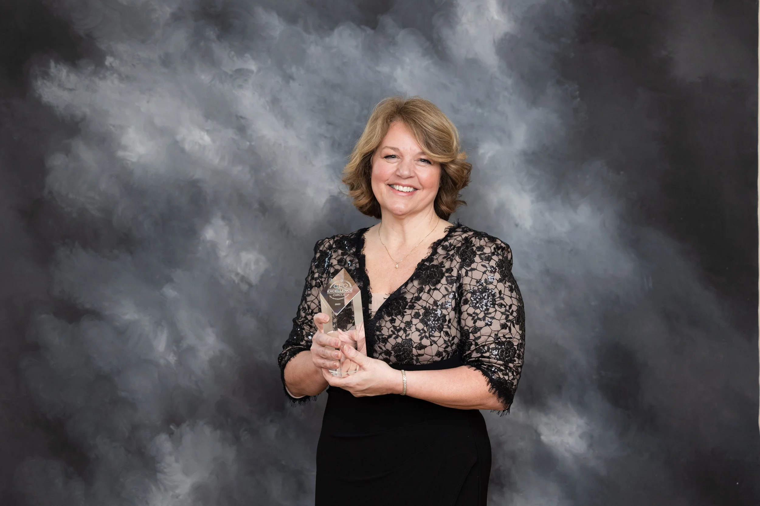 A woman in a black lace dress smiling and holding a glass award in front of a gray cloud-like background.