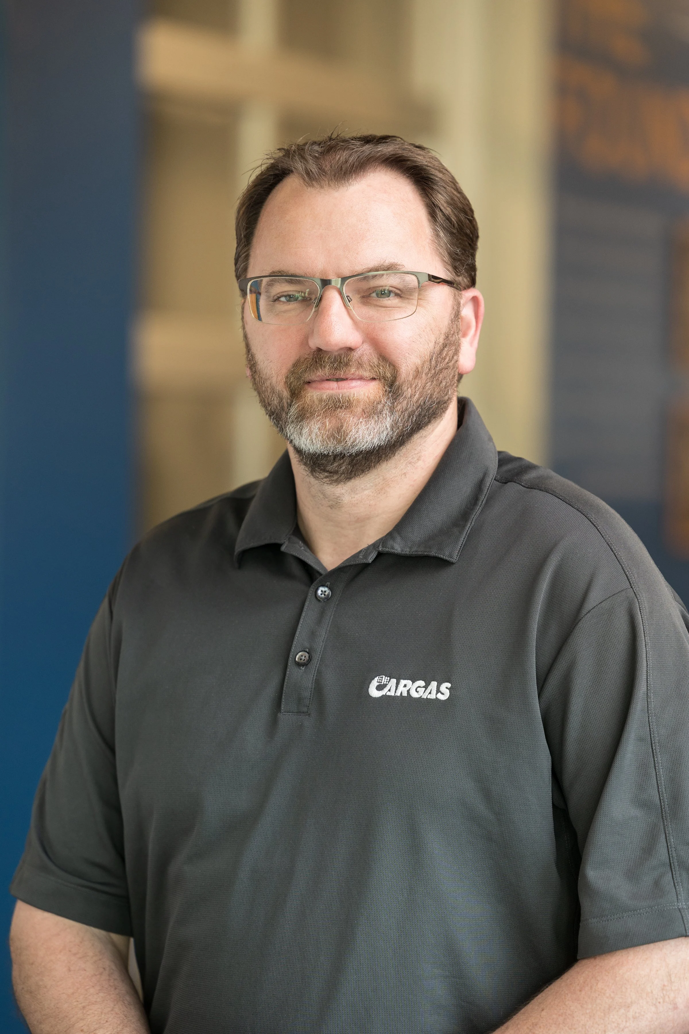 A man with glasses and a beard wearing a dark gray polo shirt with a company logo, standing in front of a blurred background of an office setting.