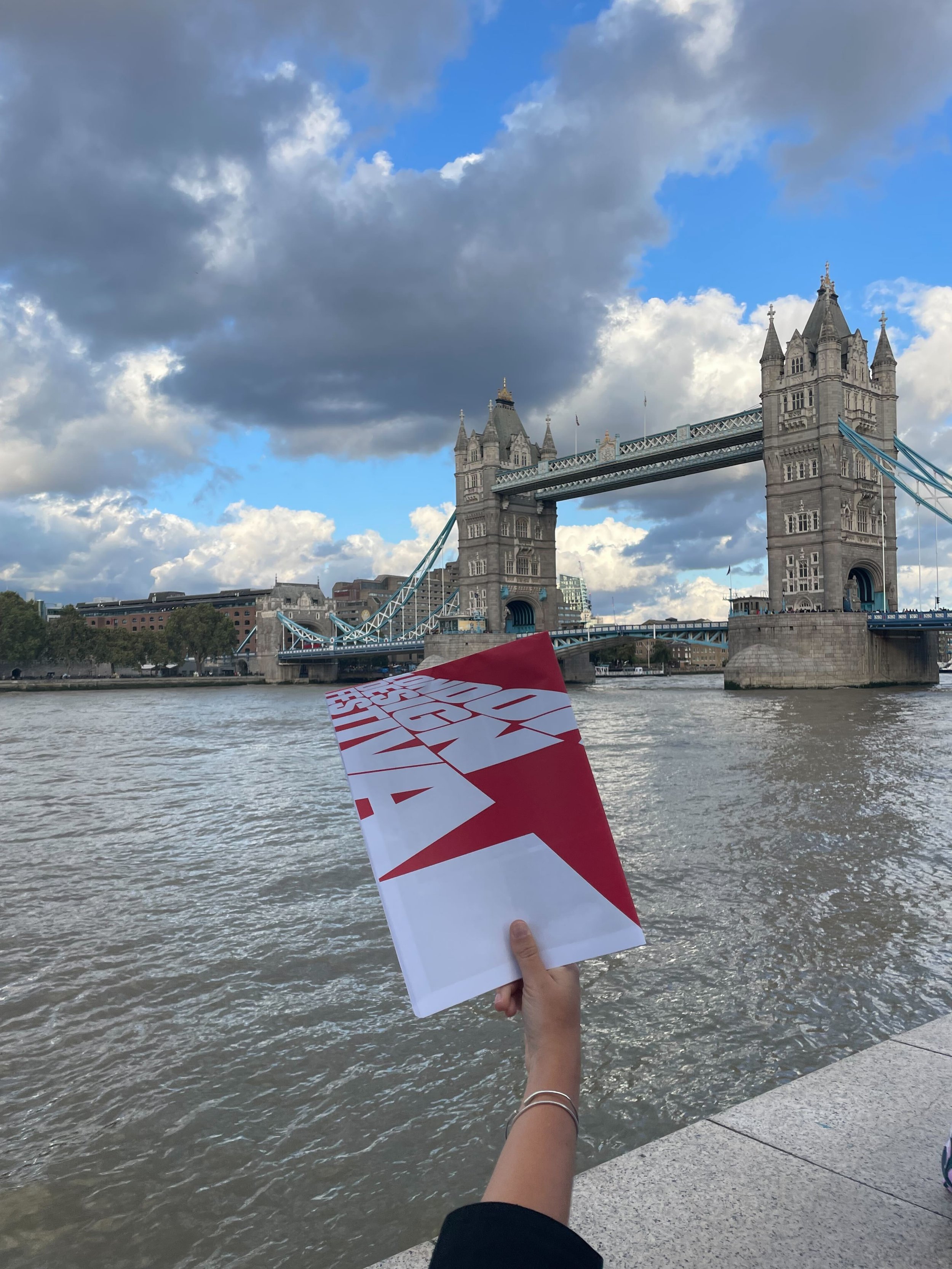  A person holding up a London Design Festival brochure in hand and the London Bridge in the background. 