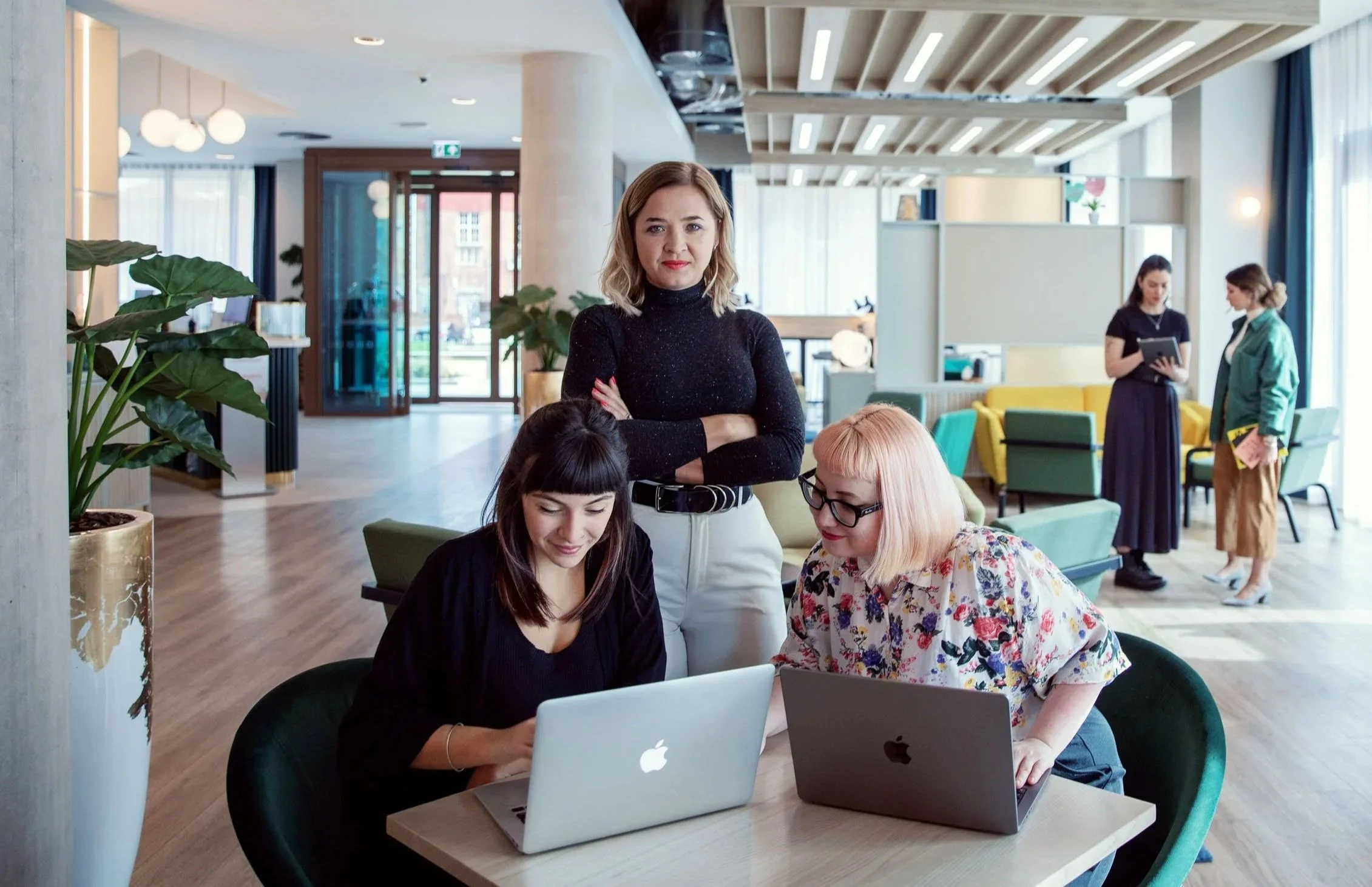 Three women working on laptops in a modern, well-lit lounge area with large windows, one woman standing behind two seated women, all engaged with their computers.