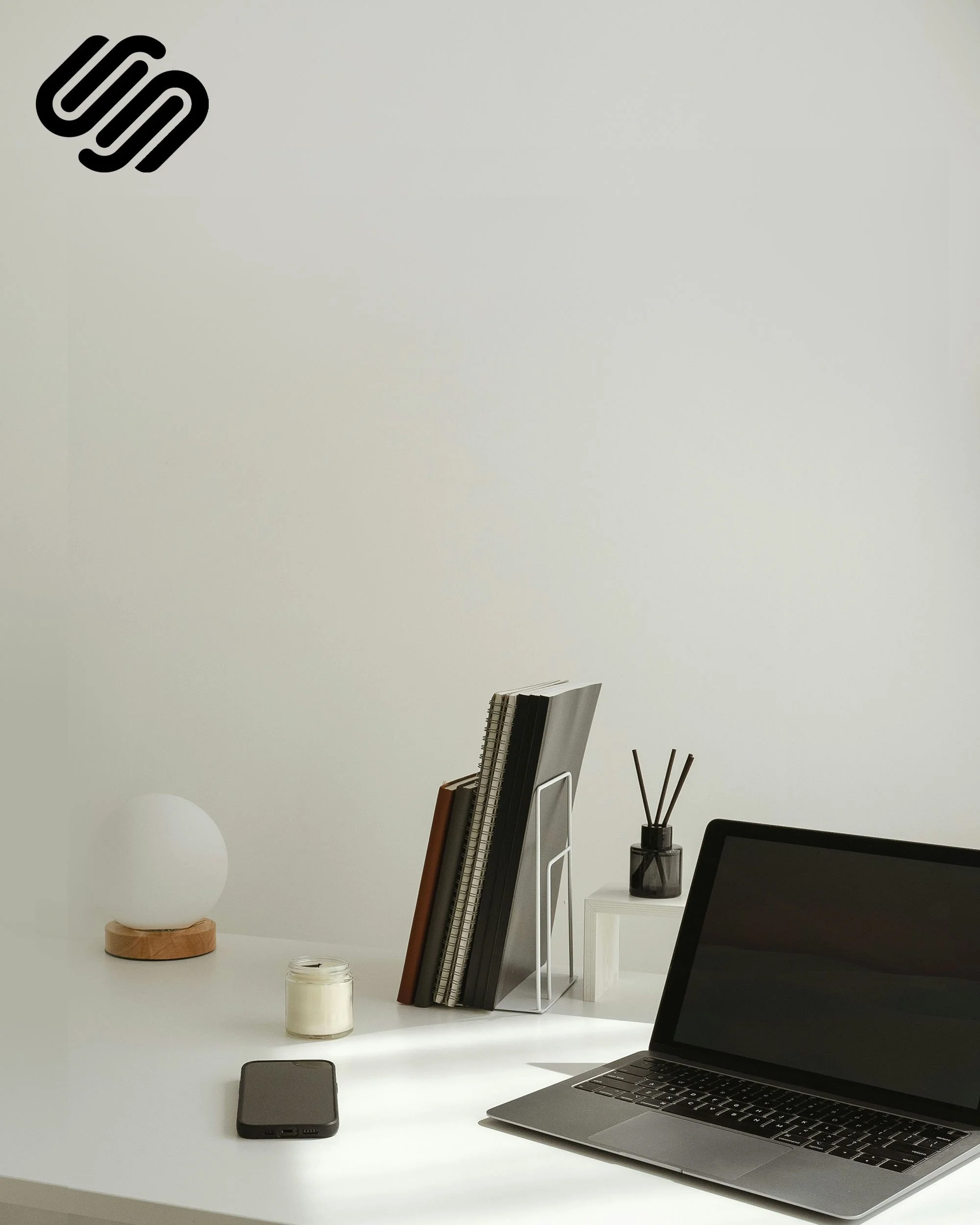 A white desk with a laptop, a black smartphone, a candle in a glass jar, a white lamp on a wooden base, and various office supplies organized in a black holder. There is a black chair in the foreground and a white side table with a black vase containing reed diffusers.