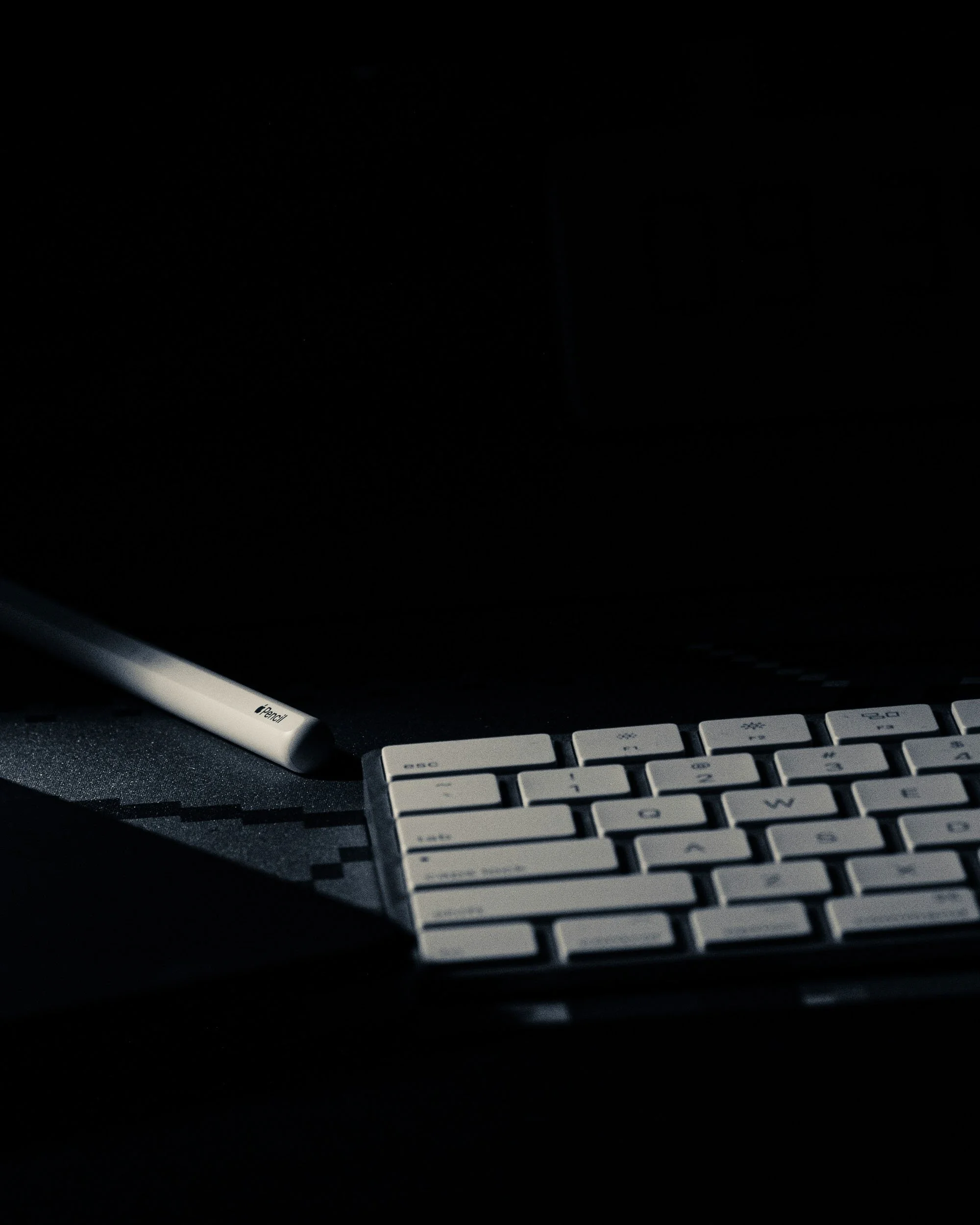 Close-up of a white Apple Pencil resting on a black desk next to a Mac keyboard, with a dark computer screen in the background.