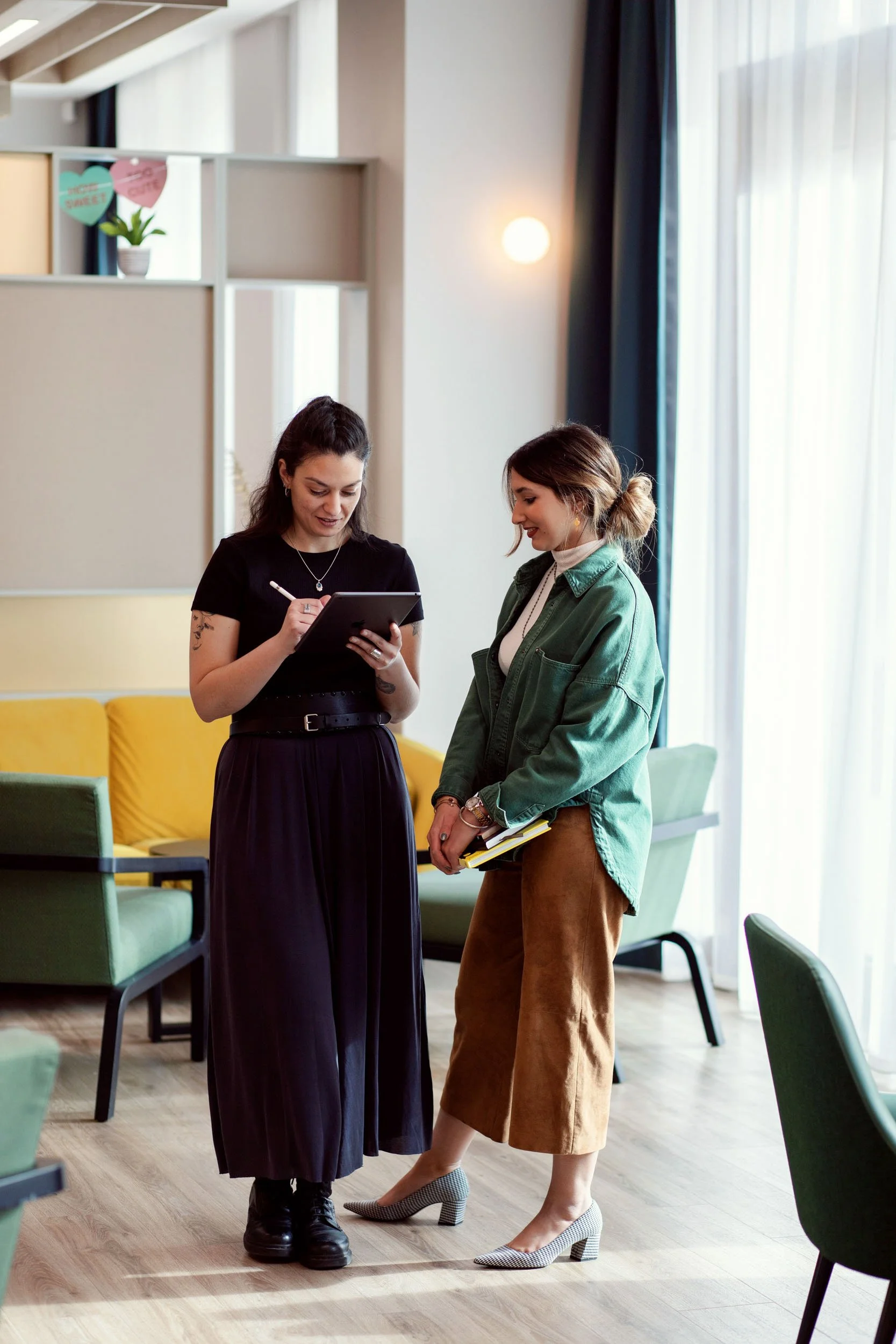 Two women standing and talking in a modern, well-lit room with colorful chairs and a yellow sofa. One woman is holding a tablet, the other is holding a notebook and both are smiling.