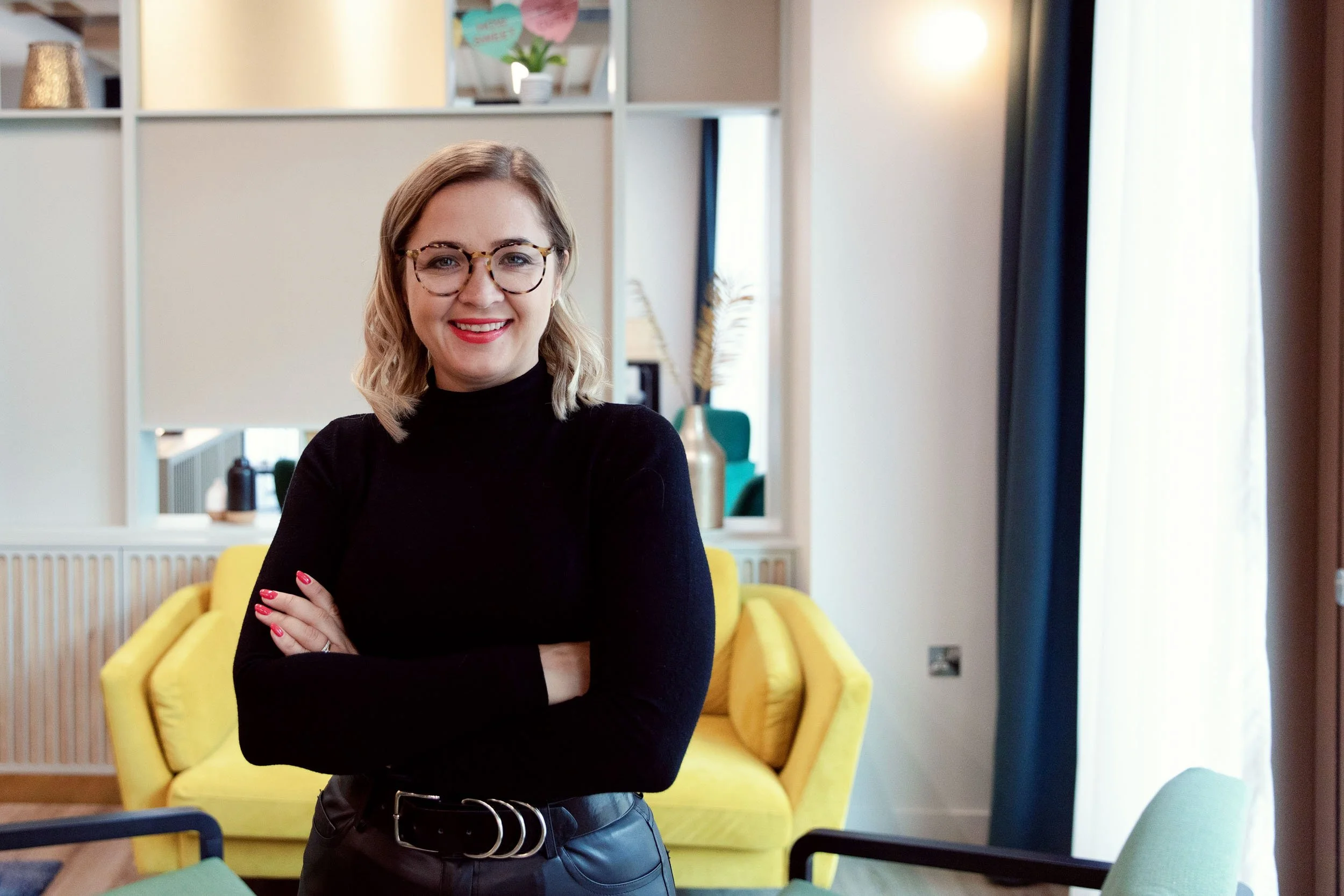A woman with blonde hair, glasses, wearing a black top, smiling and standing with crossed arms inside a modern, colorful living space with yellow chairs and decorative elements.
