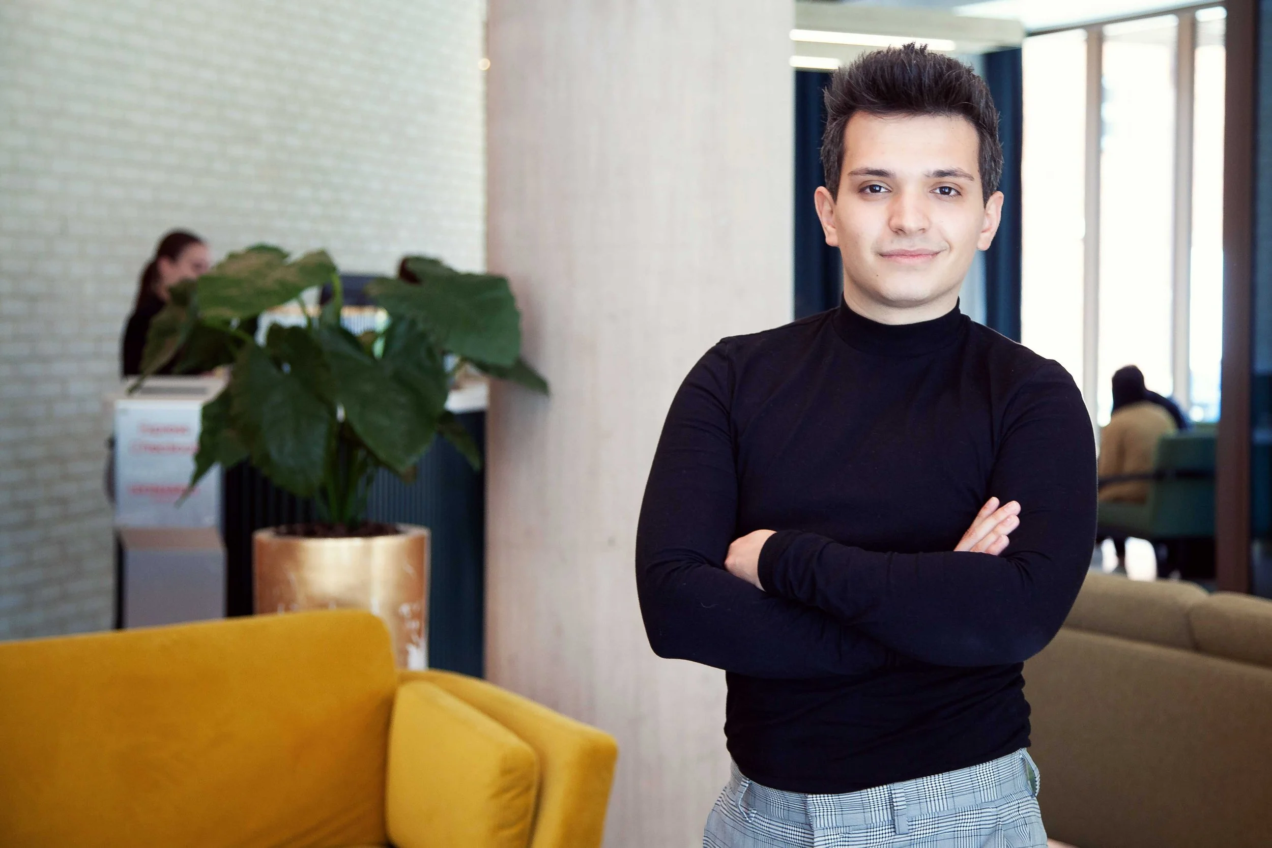 A young man with dark, styled hair and light skin stands with arms crossed in a modern indoor space, smiling subtly. He wears a black turtleneck and plaid pants. Background includes a yellow sofa, a large potted plant, and a person working at a computer.