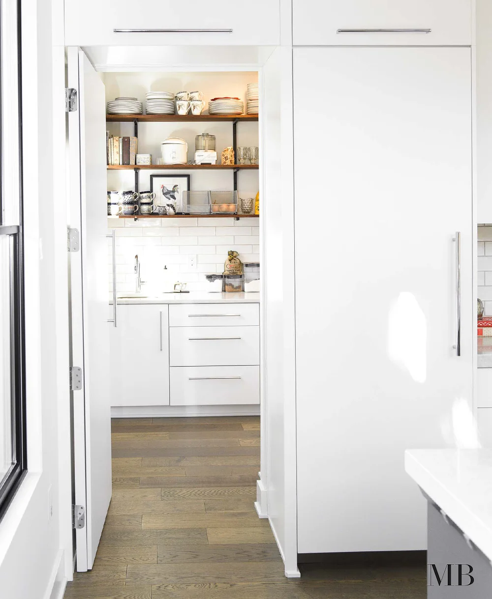 This sleek and modern kitchen remains as functional as a more traditional space by this walk-in hidden pantry. While the main space stays minimal and clear of too many objects, the pantry is full of useful kitchen objects at an arms length. This home was designed with a modern farmhouse style and we kept that going with subway tile and wooden shelves.