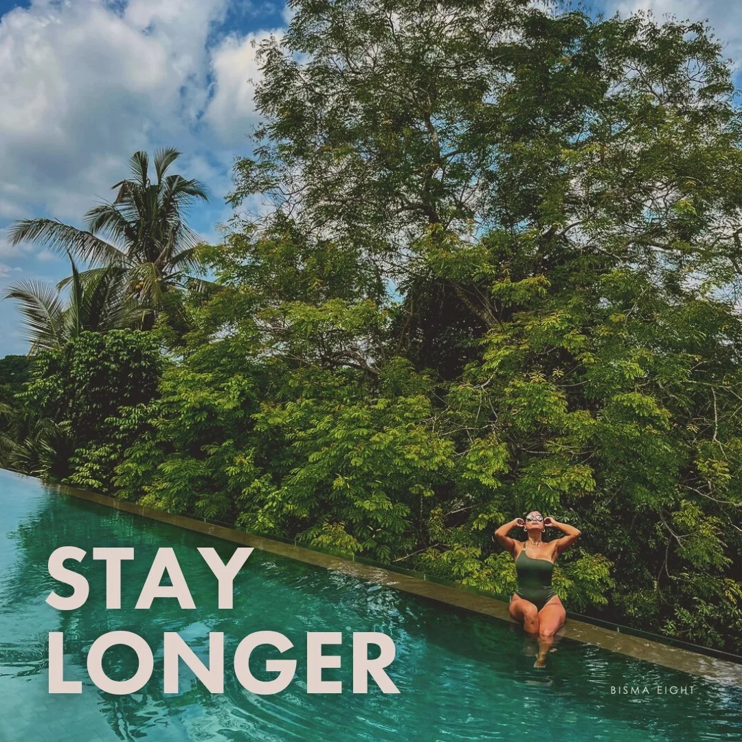 A woman wearing sunglasses and a swimsuit is sitting at the edge of a swimming pool with lush green trees in the background. The words 'STAY LONGER' are overlaid on the image.