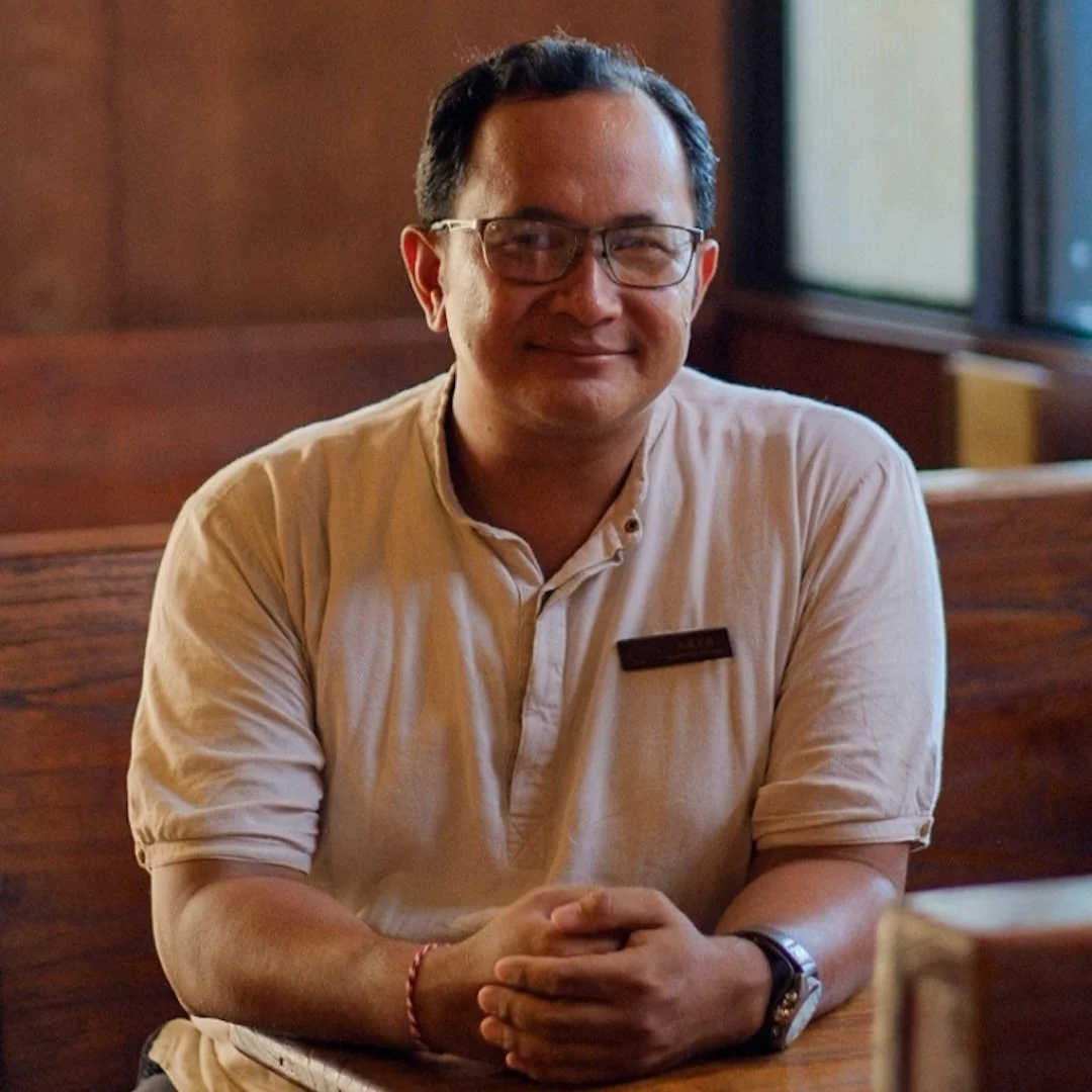 A man with glasses wearing a beige short-sleeve shirt sitting at a wooden table in a restaurant.