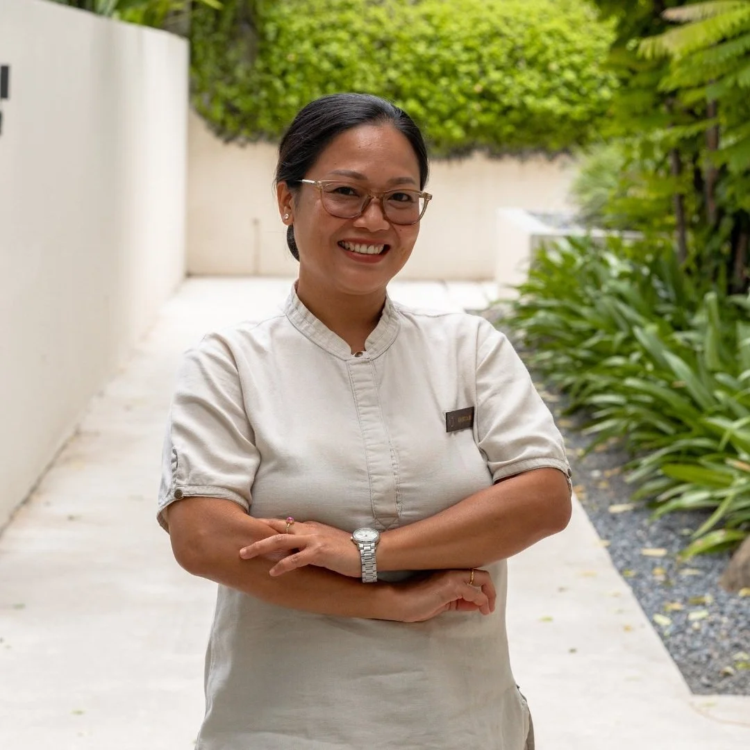 A smiling woman with glasses and a beige uniform standing outdoors with her arms crossed in front of green foliage.