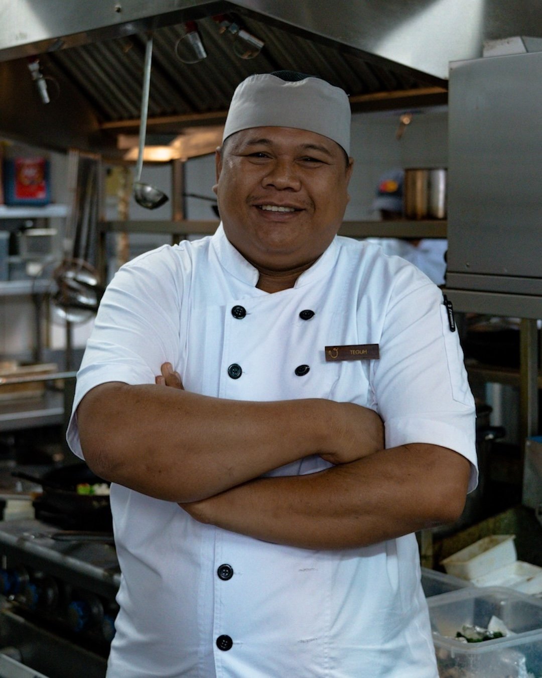 A smiling chef wearing a white chef's coat and a gray chef's hat standing with arms crossed inside a professional kitchen.