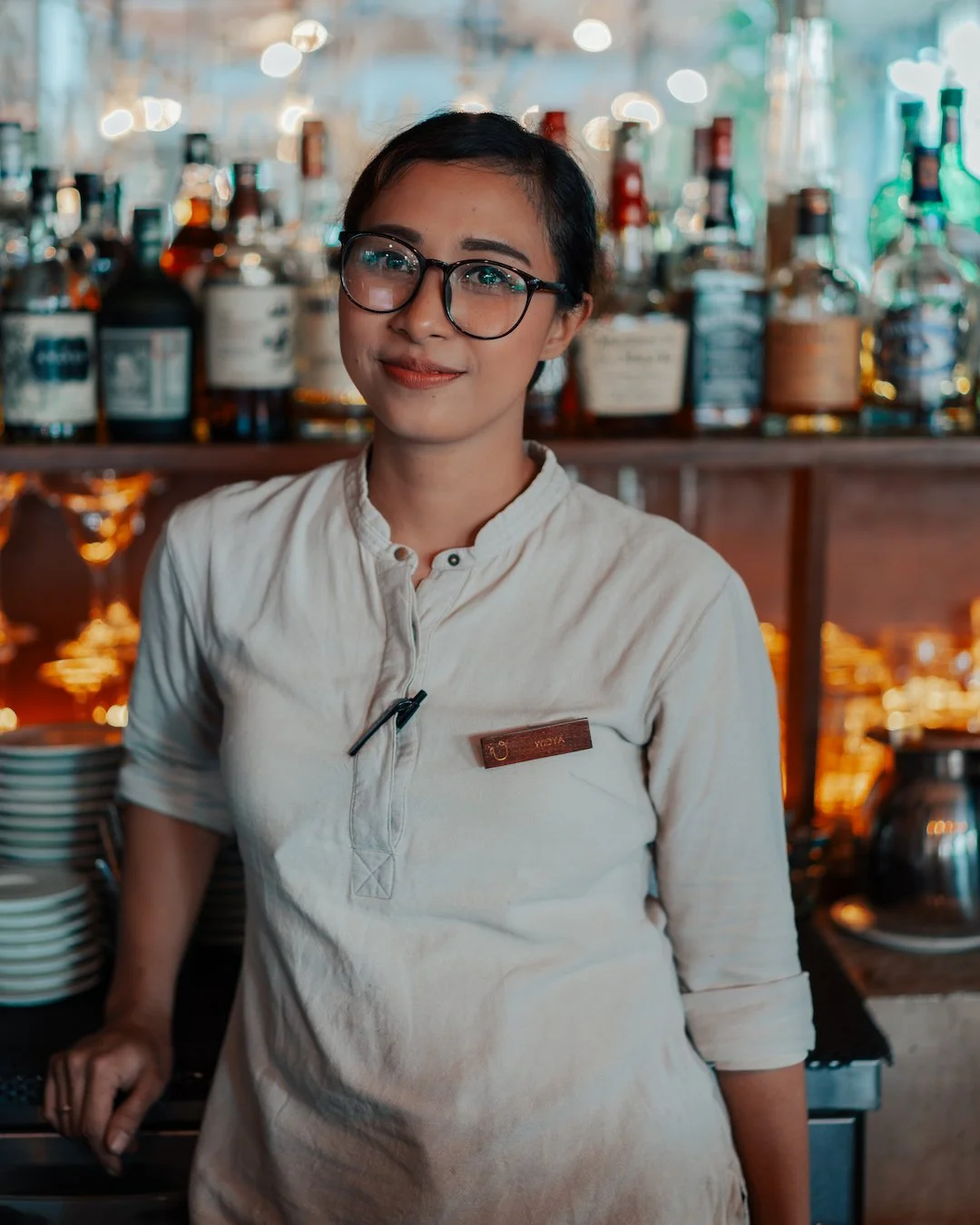 A smiling woman with glasses and a white shirt standing behind a bar counter in a dimly lit bar or restaurant, with bottles of alcohol on shelves behind her.