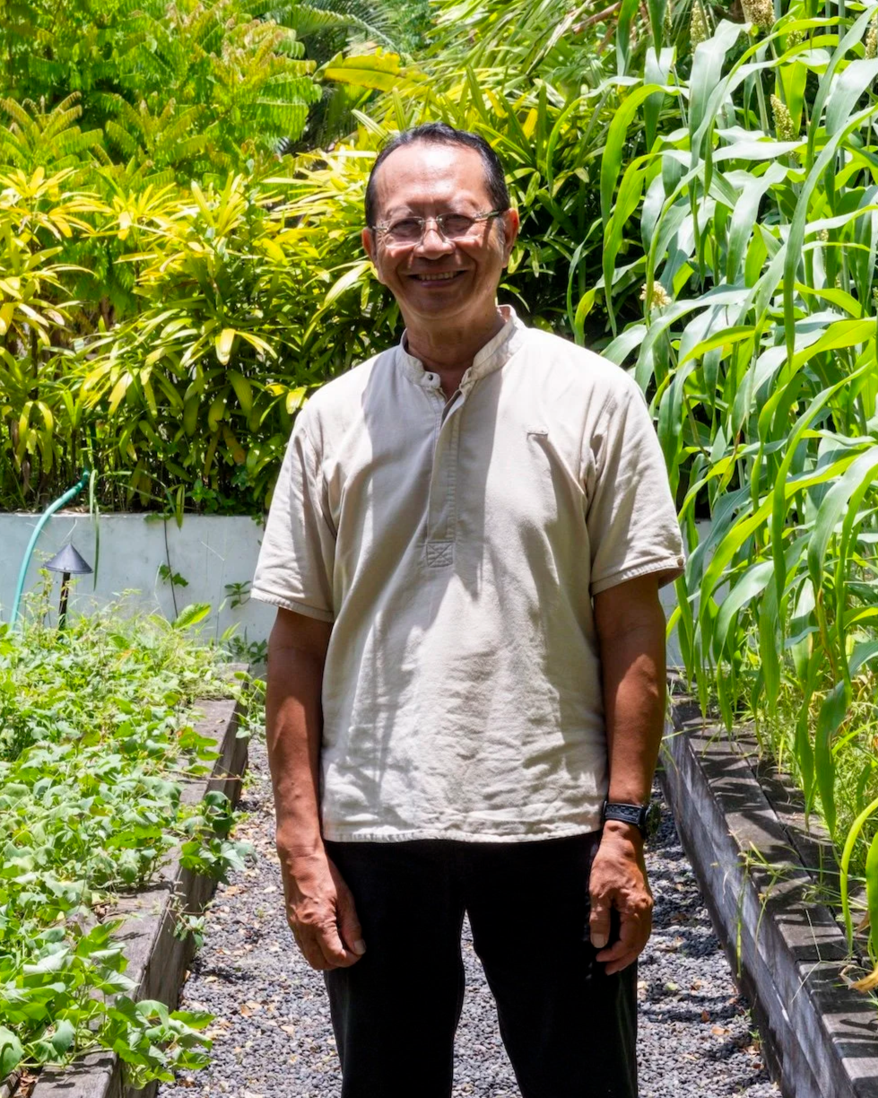 A smiling middle-aged man in a beige shirt standing in a lush green garden with tall plants and a vegetable bed.