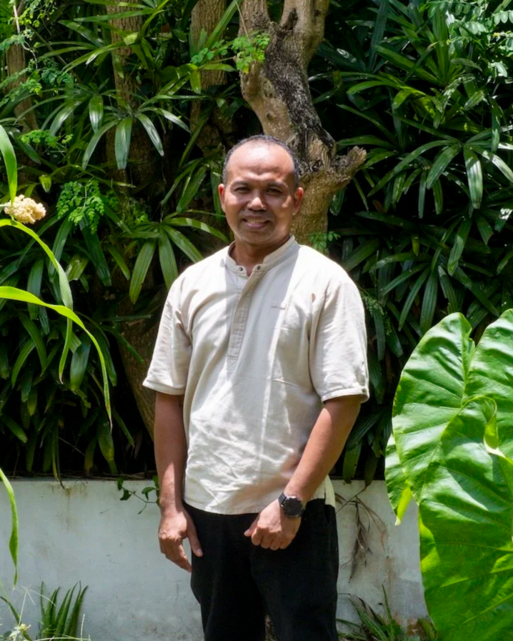 A man with a shaved head wearing a beige traditional shirt and black pants, standing outdoors in front of lush green tropical plants and a tree.