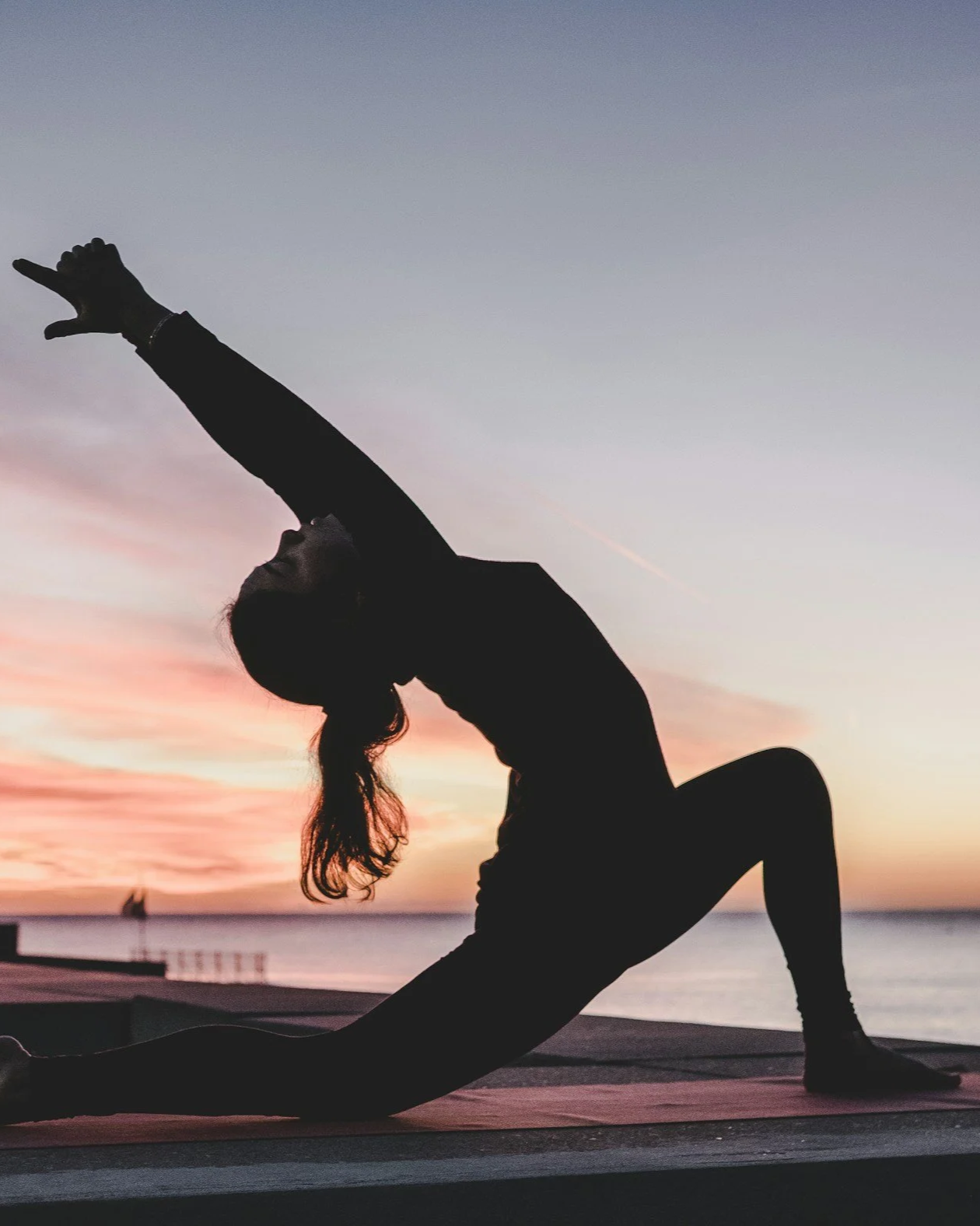 Silhouette of a woman practicing yoga outdoors at sunset, performing a pose with one arm extended upward and the other arm supporting her on the ground, near a body of water.