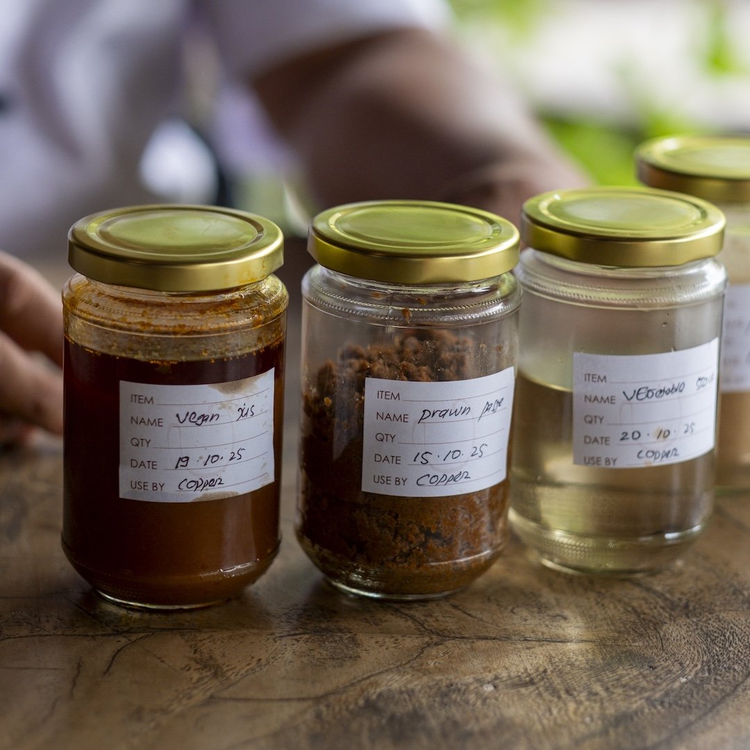 Three glass jars with gold lids containing various food items are placed on a wooden surface. Each jar has a handwritten label with item names, quantities, dates, and use-by dates. The items include vegan miso, dried prawn, and vegetable oil.