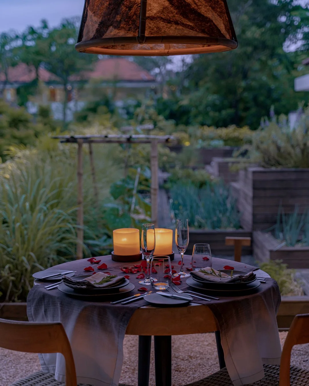 A romantic dinner at Embers with a white tablecloth set for two, decorated with red rose petals and napkins folded with sprigs of herbs. Inside a cozy, outdoor garden area with greenery and wooden planters, illuminated by warm lighting.