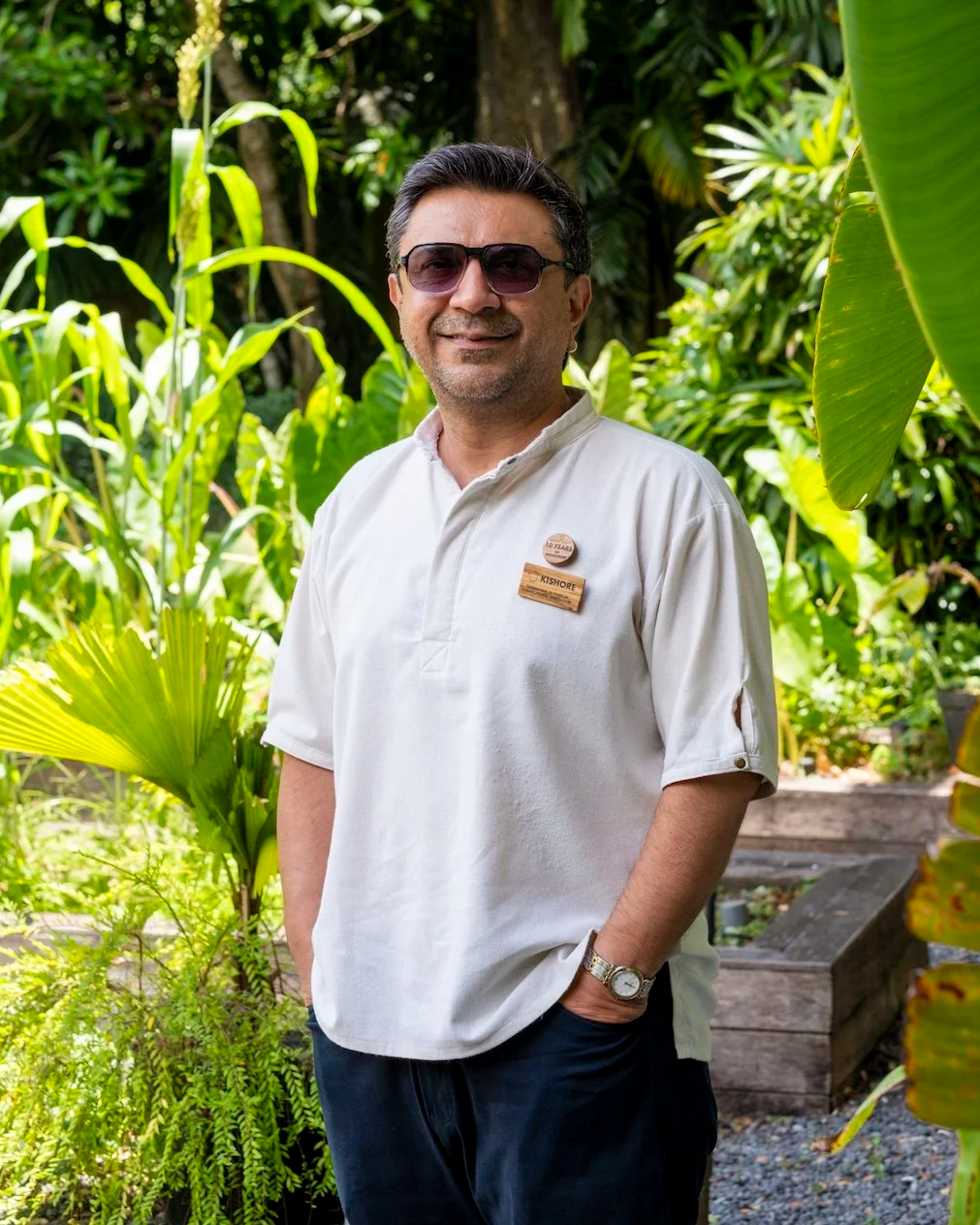 A man with dark hair, wearing sunglasses, a white polo shirt, and a watch, standing outdoors among green tropical plants.