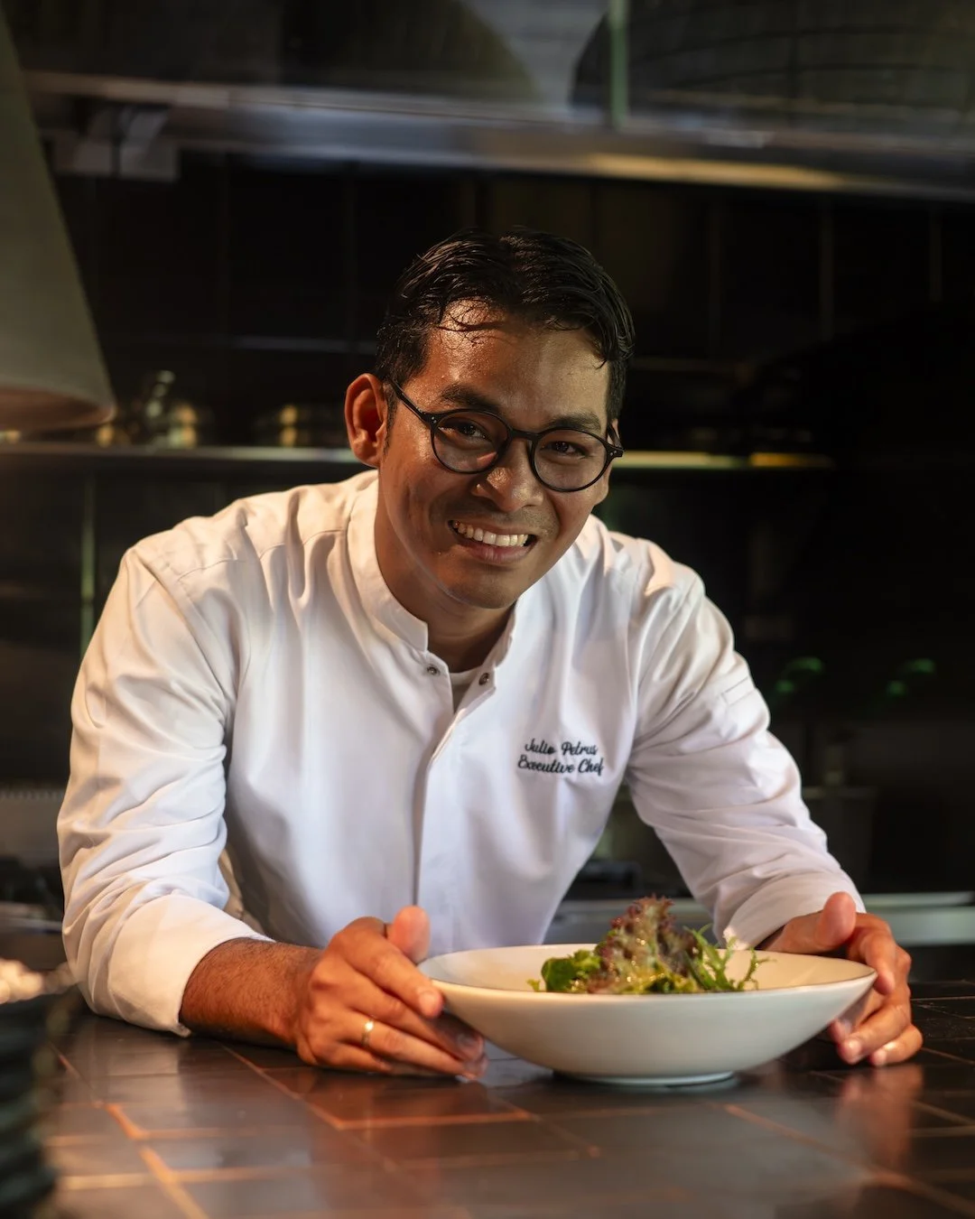 A smiling chef in a white uniform and glasses presenting a bowl of salad in a restaurant kitchen.