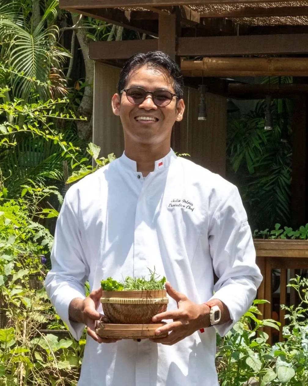 A smiling man in a white chef's uniform and sunglasses holding a bowl of greens outdoors.