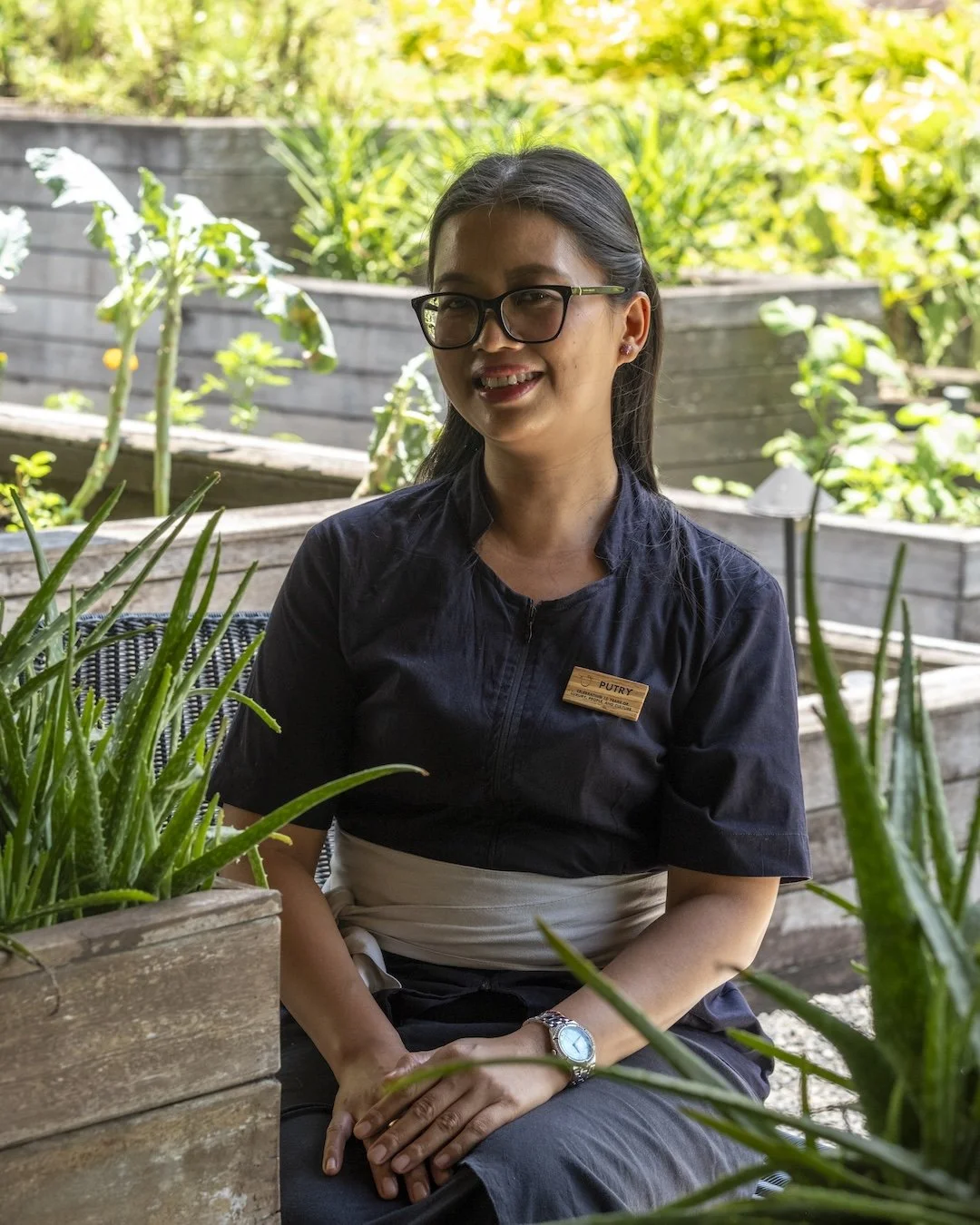A woman with glasses and a name tag sitting outdoors among wooden planters with green plants, smiling at the camera.