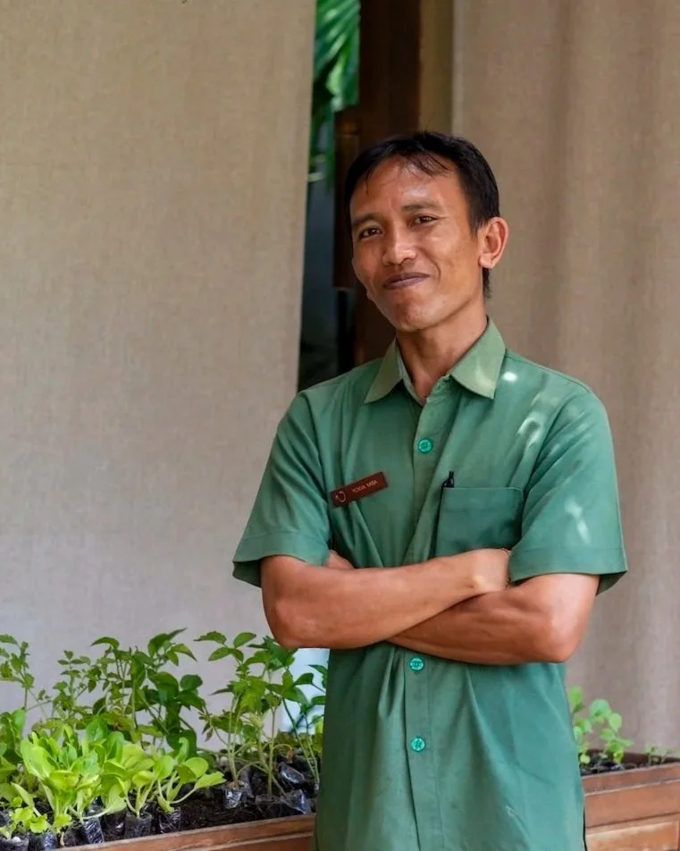 A man in a light green uniform with a name tag stands with arms crossed, smiling gently, in front of a wall and some potted plants.