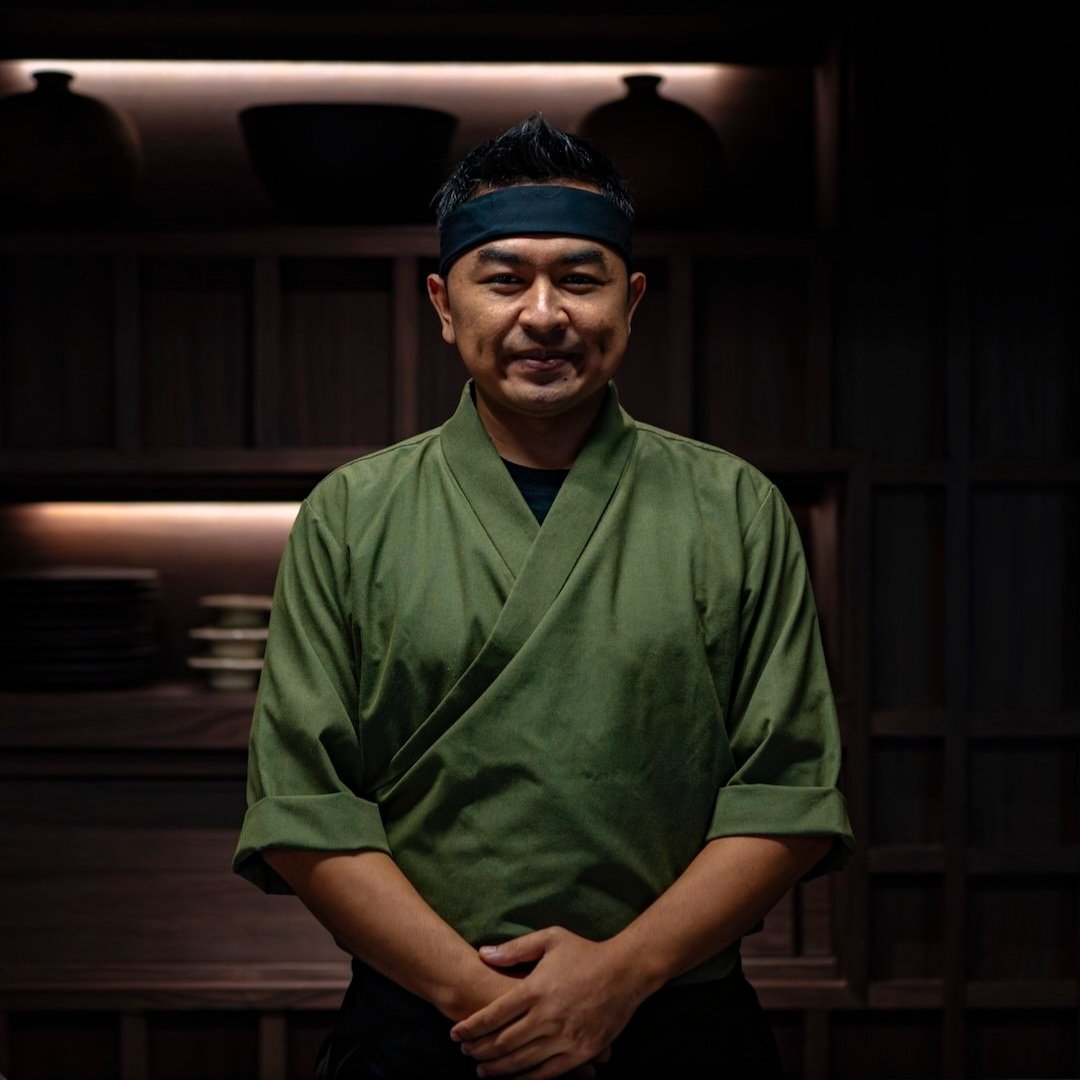 A man wearing a green traditional Japanese chef's uniform and a black headband, standing in a dark kitchen or restaurant setting.