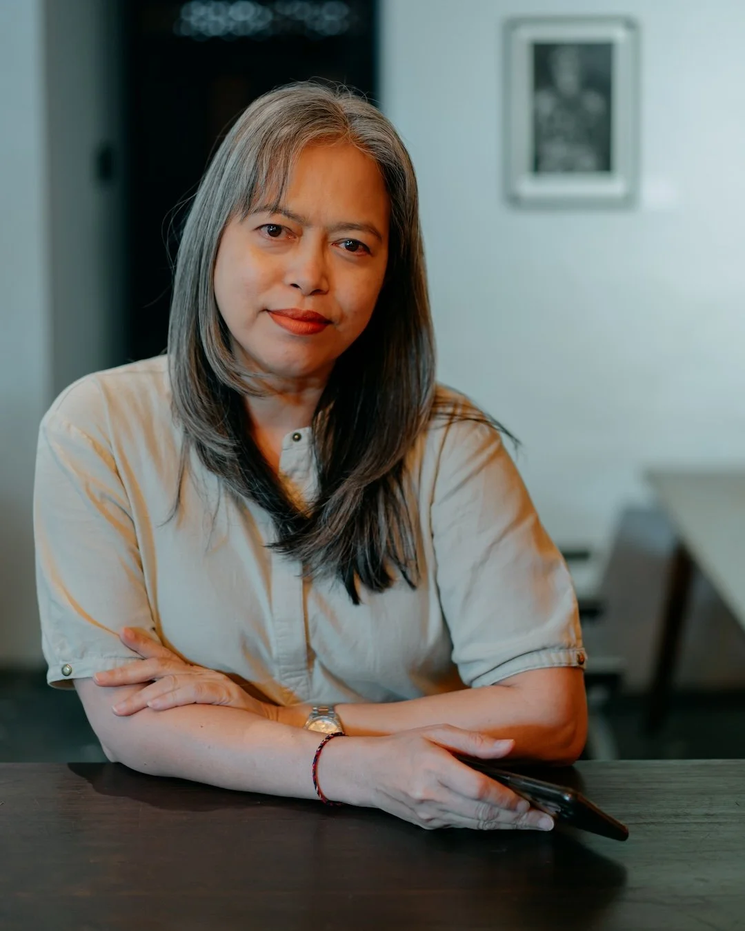 A woman with shoulder-length dark hair and light skin sitting at a dark wooden table, holding a smartphone, looking at the camera, in a room with framed photo on the wall in the background.