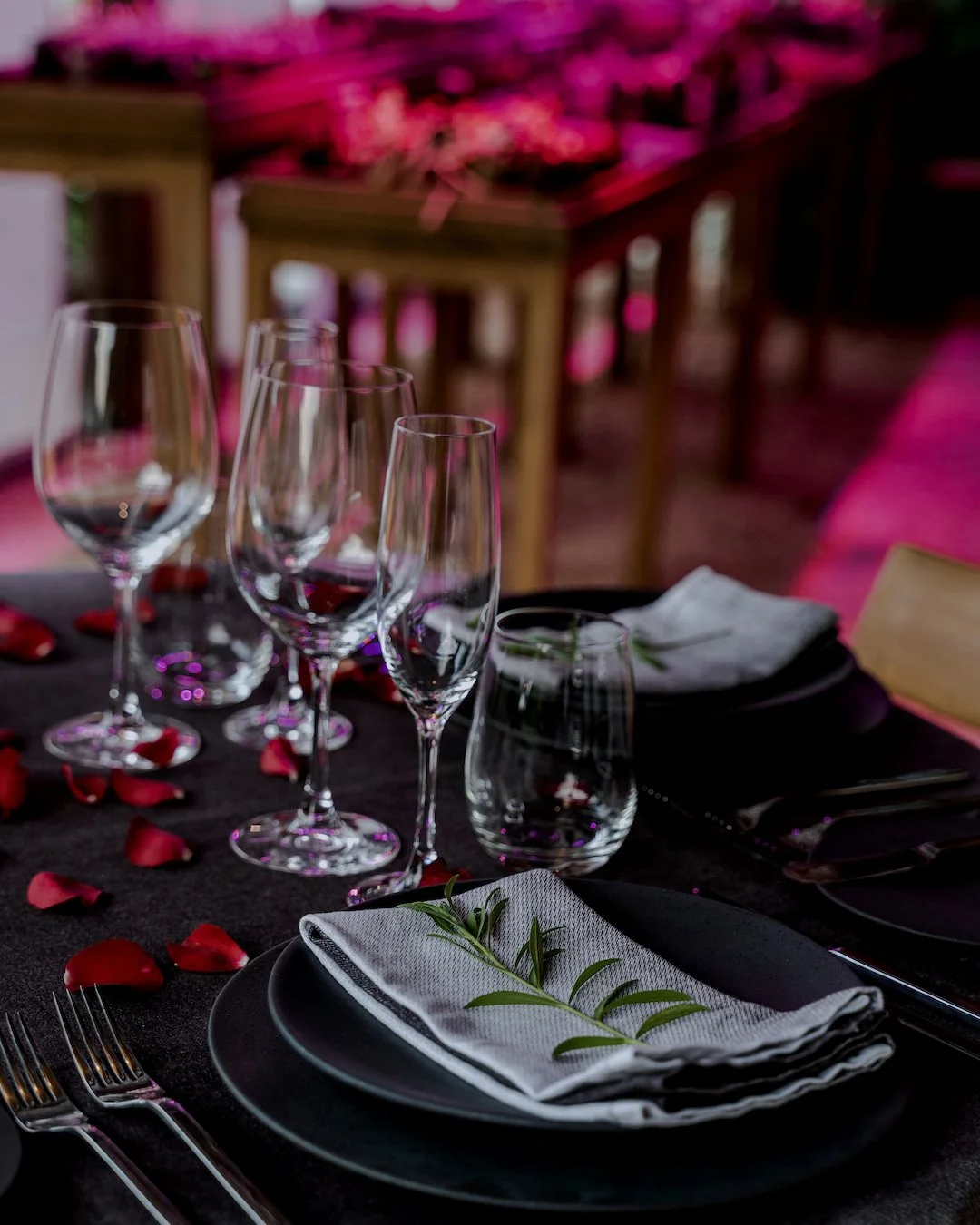 A romantic dinner at Embers, table setting with wine glasses, water glasses, black plates, white napkins with a green leaf, and scattered rose petals, in a restaurant with pink lighting.