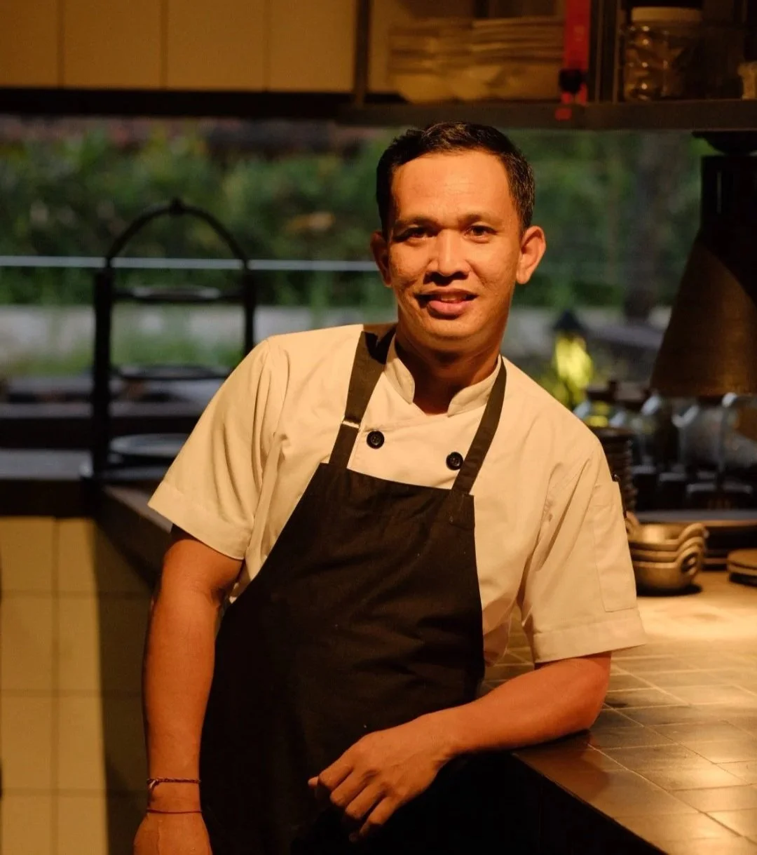 Embers Sous Chef in a beige chef coat and black apron standing behind a Embers Ubud kitchen counter, smiling at the camera.