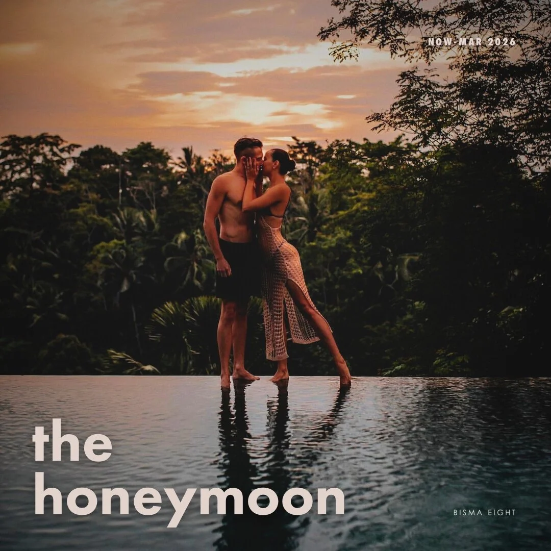 A man and woman are kissing at the edge of a pool against a sunset sky with lush green trees in the background.